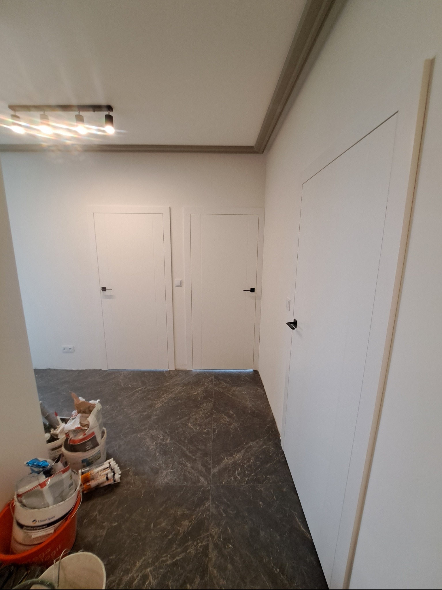 Białe drzwi wewnętrzne z czarnymi klamkami w wykończonym korytarzu z ciemną podłogą. Widoczne materiały budowlane w rogu pomieszczenia.
