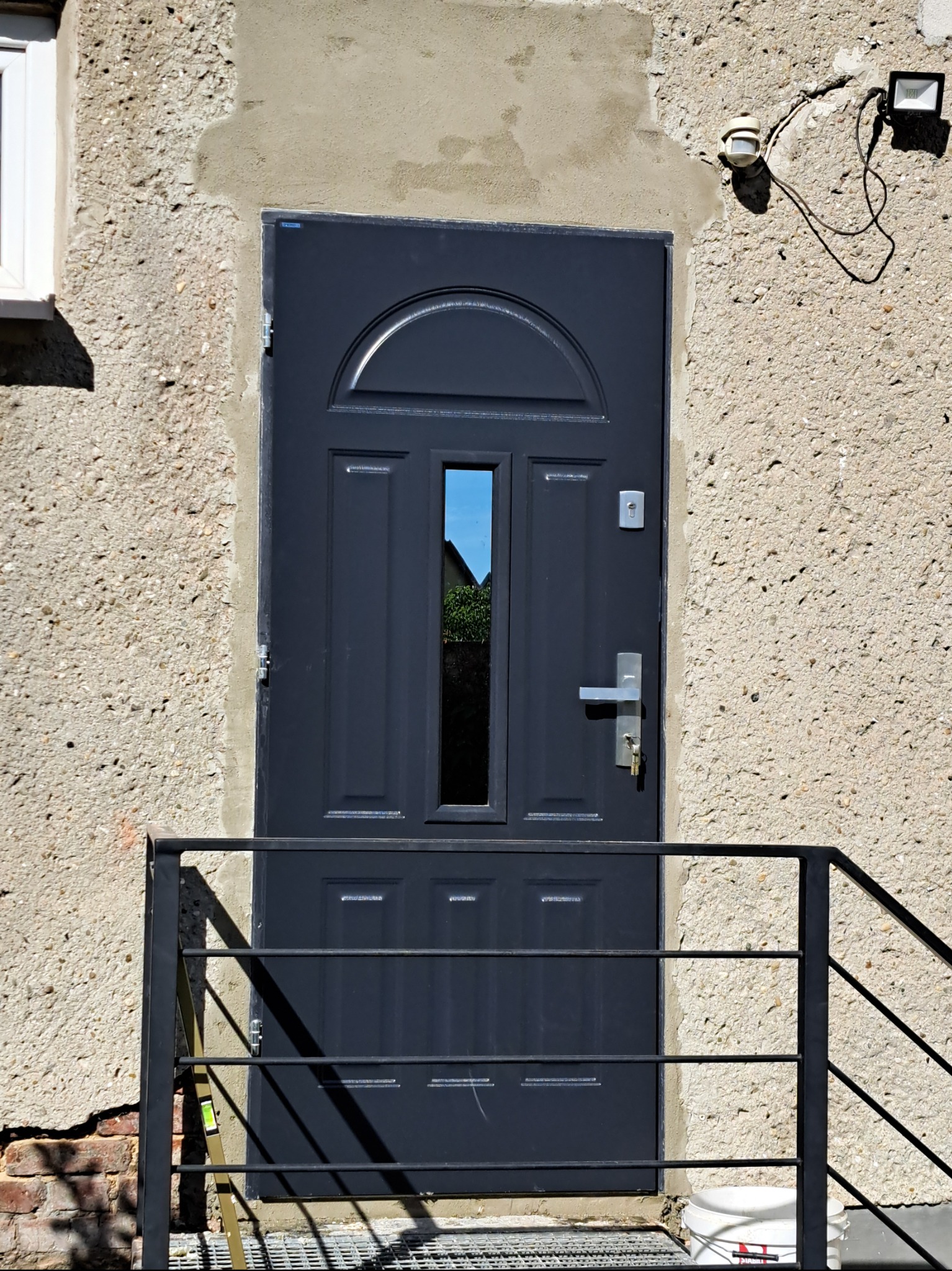 Szare drzwi wejściowe z szybką, osadzone w surowej elewacji, z widocznymi śladami prac wykończeniowych i metalową barierką. Widoczny poziomica.