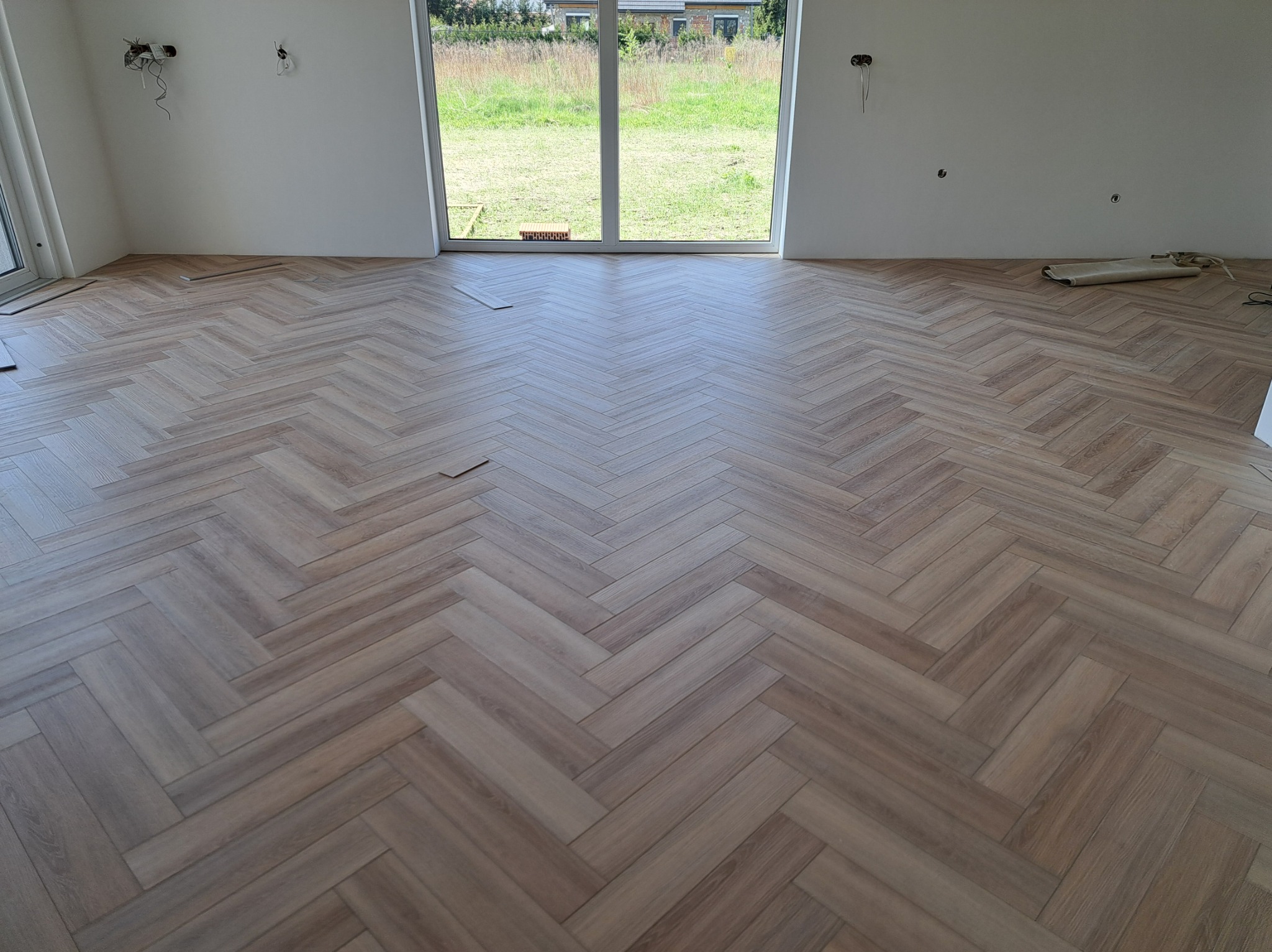 Ukończona podłoga z paneli ułożonych w jodełkę w jasnym, przestronnym wnętrzu z dużym oknem i widokiem na zieleń. Widoczne niedokończone ściany i instalacje.