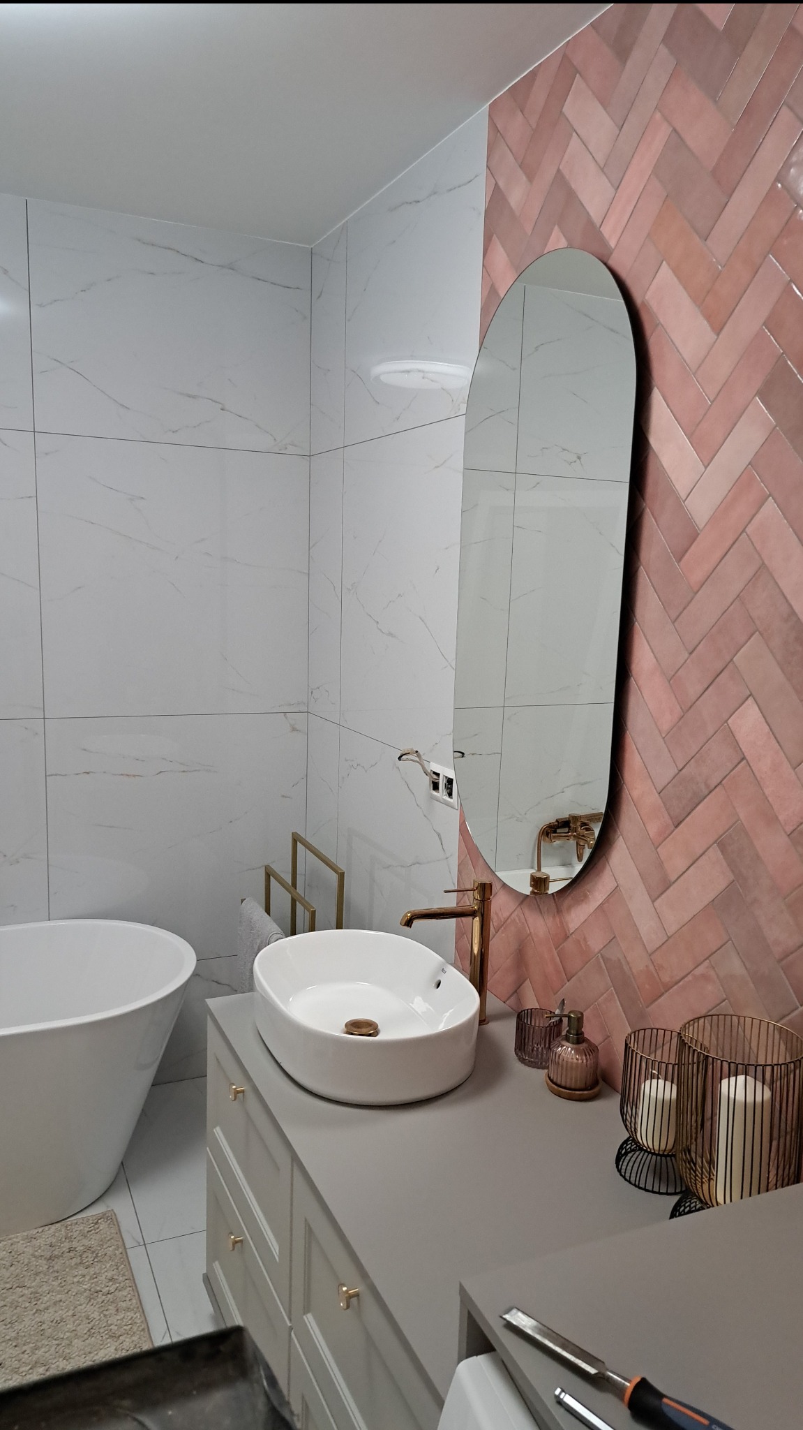 Nowoczesna łazienka z białą armaturą, różową ścianą w jodełkę i owalnym lustrem. Umywalka nablatowa, złota bateria i dekoracje dodają elegancji.
