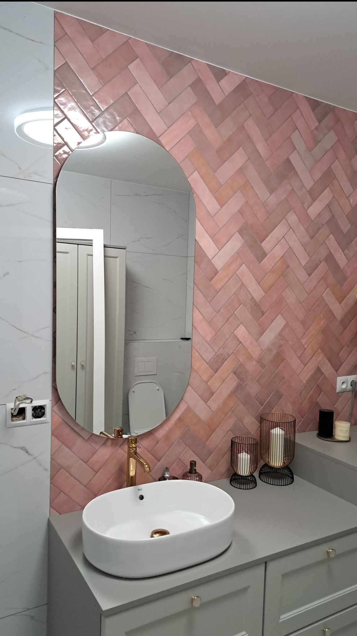 Nowoczesna łazienka z umywalką nablatową, złotą armaturą i różową ścianą z płytek ułożonych w jodełkę. W lustrze odbija się toaleta i szafka. Widoczne gniazdko elektryczne.