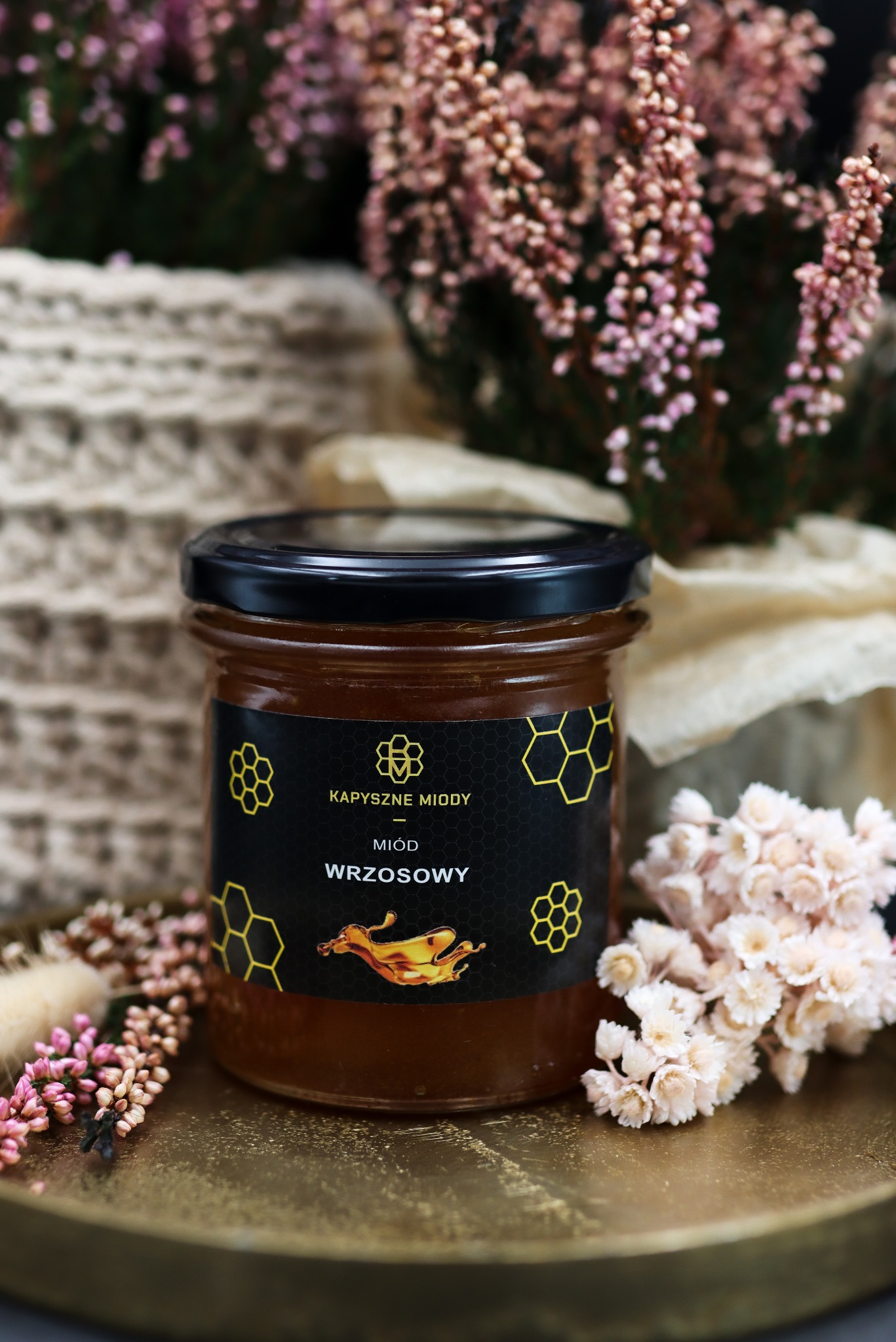 Słoik miodu wrzosowego marki Kapyszne Miody na złotej tacy, otoczony wrzosami i dekoracyjną tkaniną. Etykieta czarna z motywem plastra miodu i kroplą miodu.