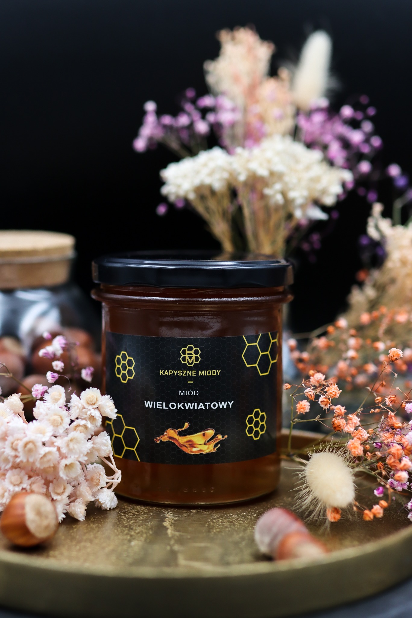 Słoik miodu wielokwiatowego marki Kapyszne Miody na złotej tacy, otoczony suszonymi kwiatami. Etykieta z motywem plastra miodu i kroplą miodu.