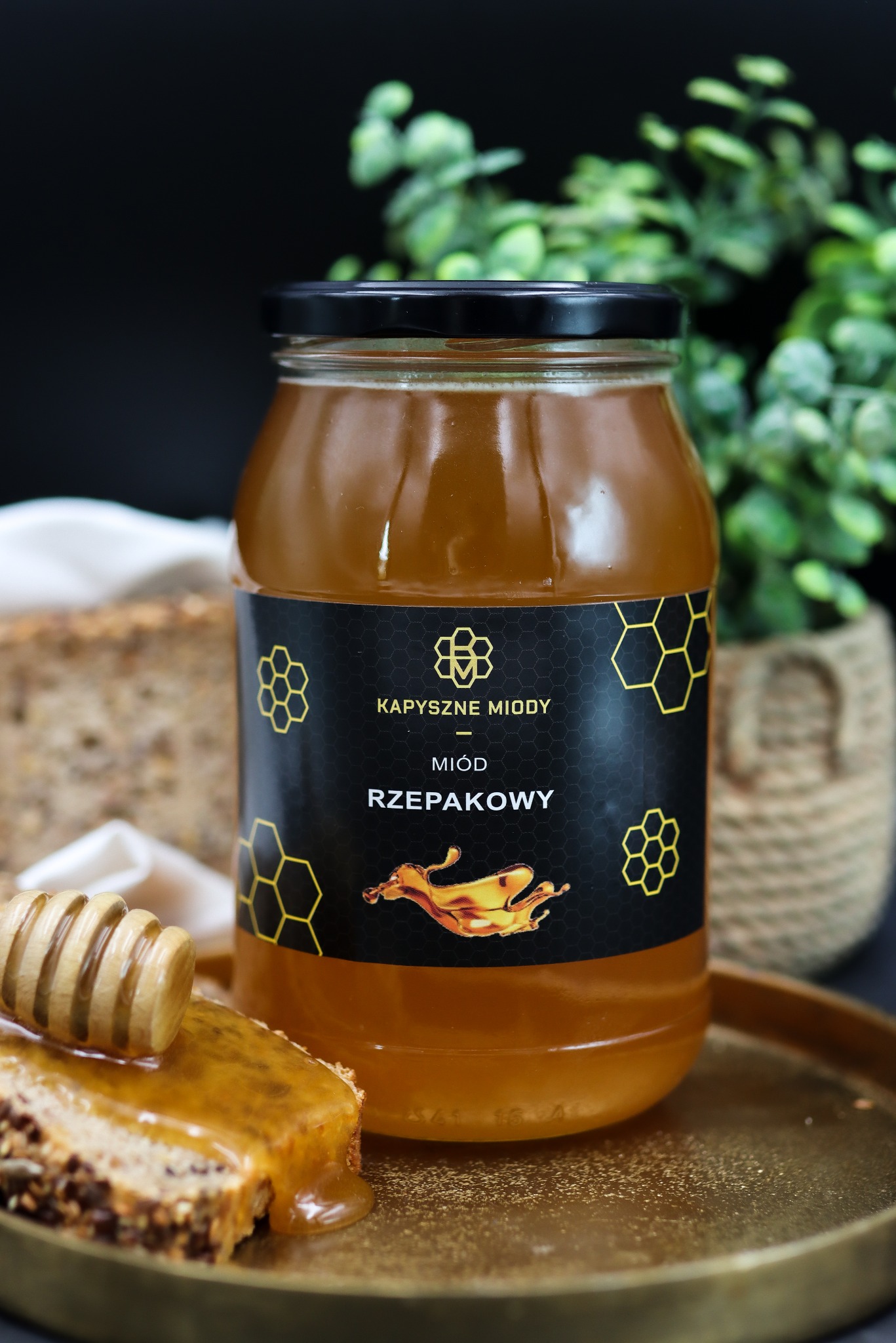 Słoik miodu rzepakowego marki Kapyszne Miody na złotym talerzu, obok kromka chleba z miodem i nabierak. W tle roślina doniczkowa i ciemne tło.
