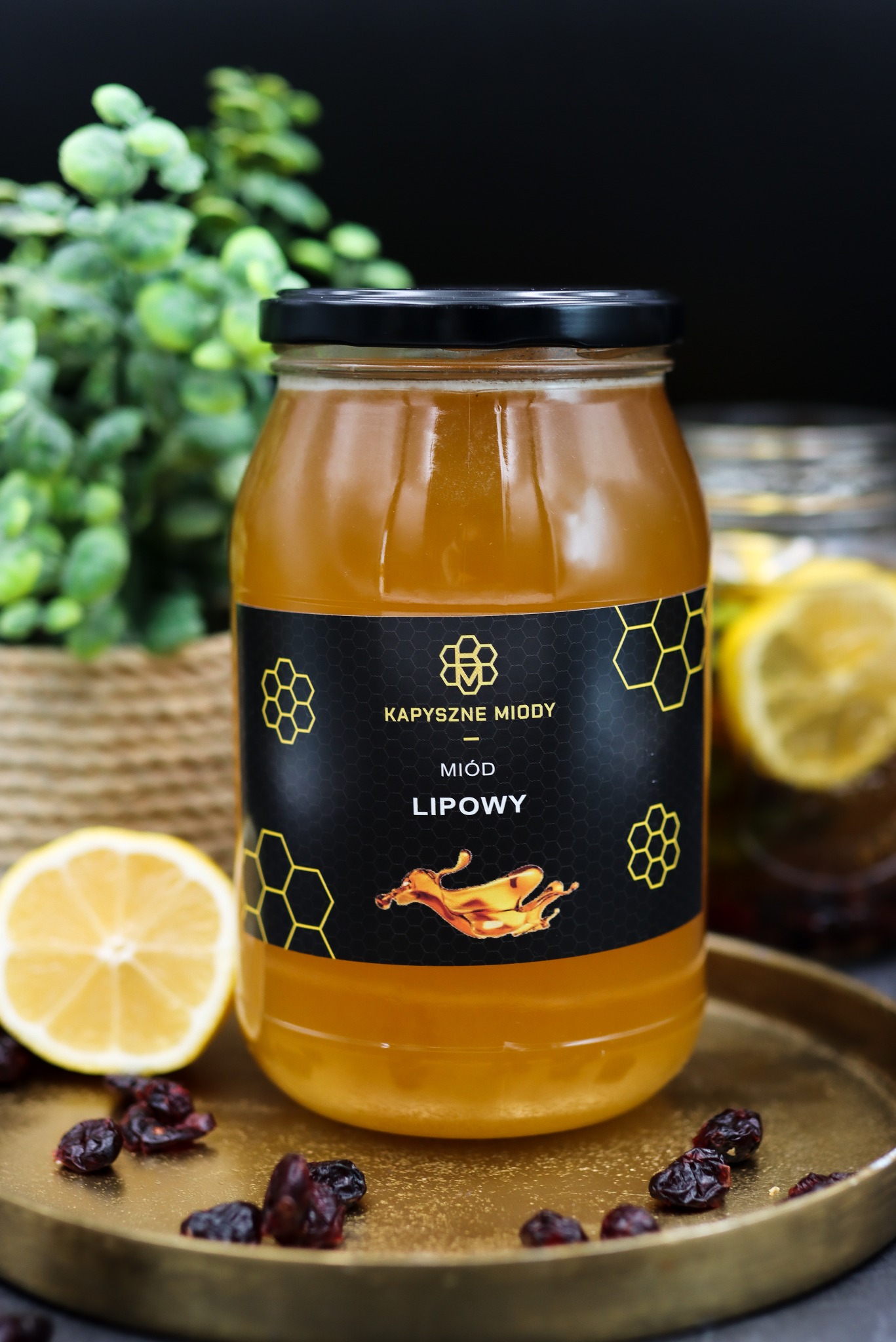 Słoik lipowego przysmaku 'Kapyszne Miody' na złotej tacy z cytryną i żurawiną. W tle zielona roślina i słoik z napojem cytrynowym.
