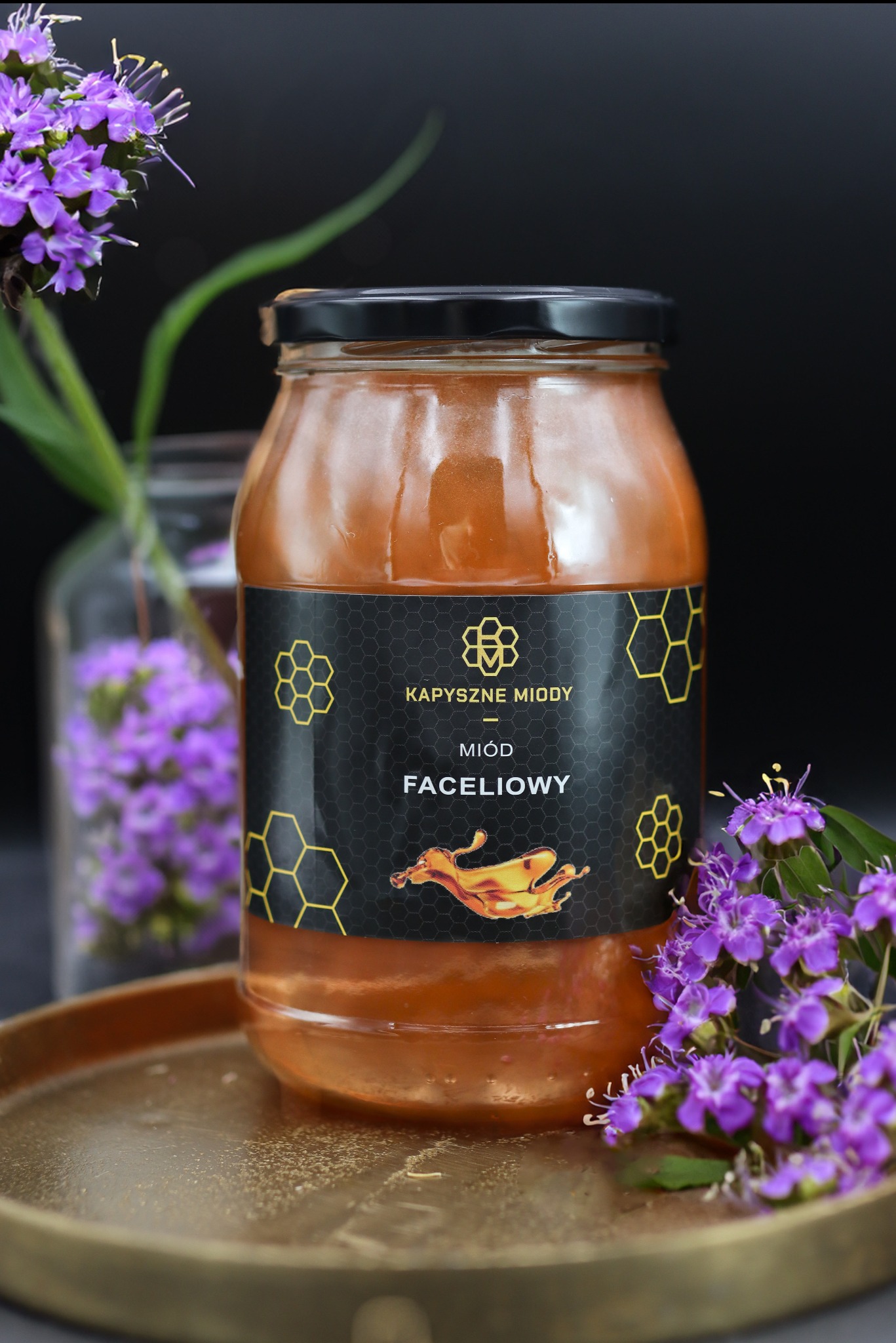 Słoik miodu faceliowego z czarną etykietą i grafiką plastra miodu, otoczony fioletowymi kwiatami na złotej tacy, na ciemnym tle. Elegancka prezentacja produktu.