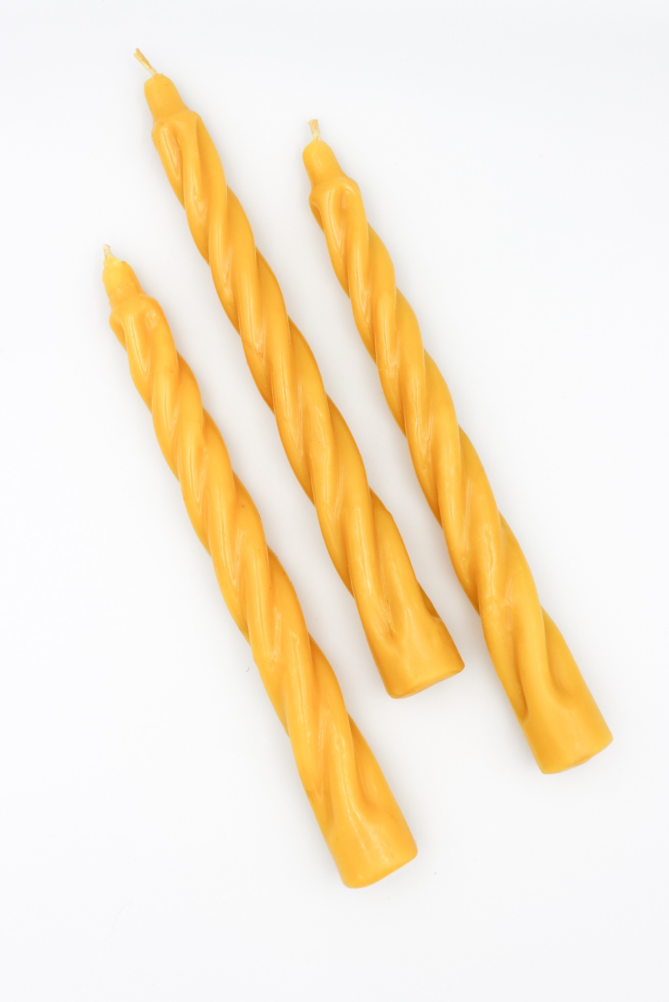 Trzy skręcone świece z wosku pszczelego w odcieniach ciepłego złota, ułożone ukośnie na białym tle. Widoczne delikatne detale skręcenia i naturalna tekstura wosku.