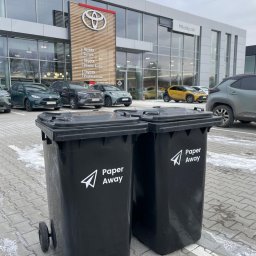 RX SPÓŁKA Z OGRANICZONĄ ODPOWIEDZIALNOŚCIĄ - Dwa czarne pojemniki na kółkach z logo Paper Away stoją przed salonem samochodowym Toyota Mikołajczak. Usługa odbioru dokumentów.