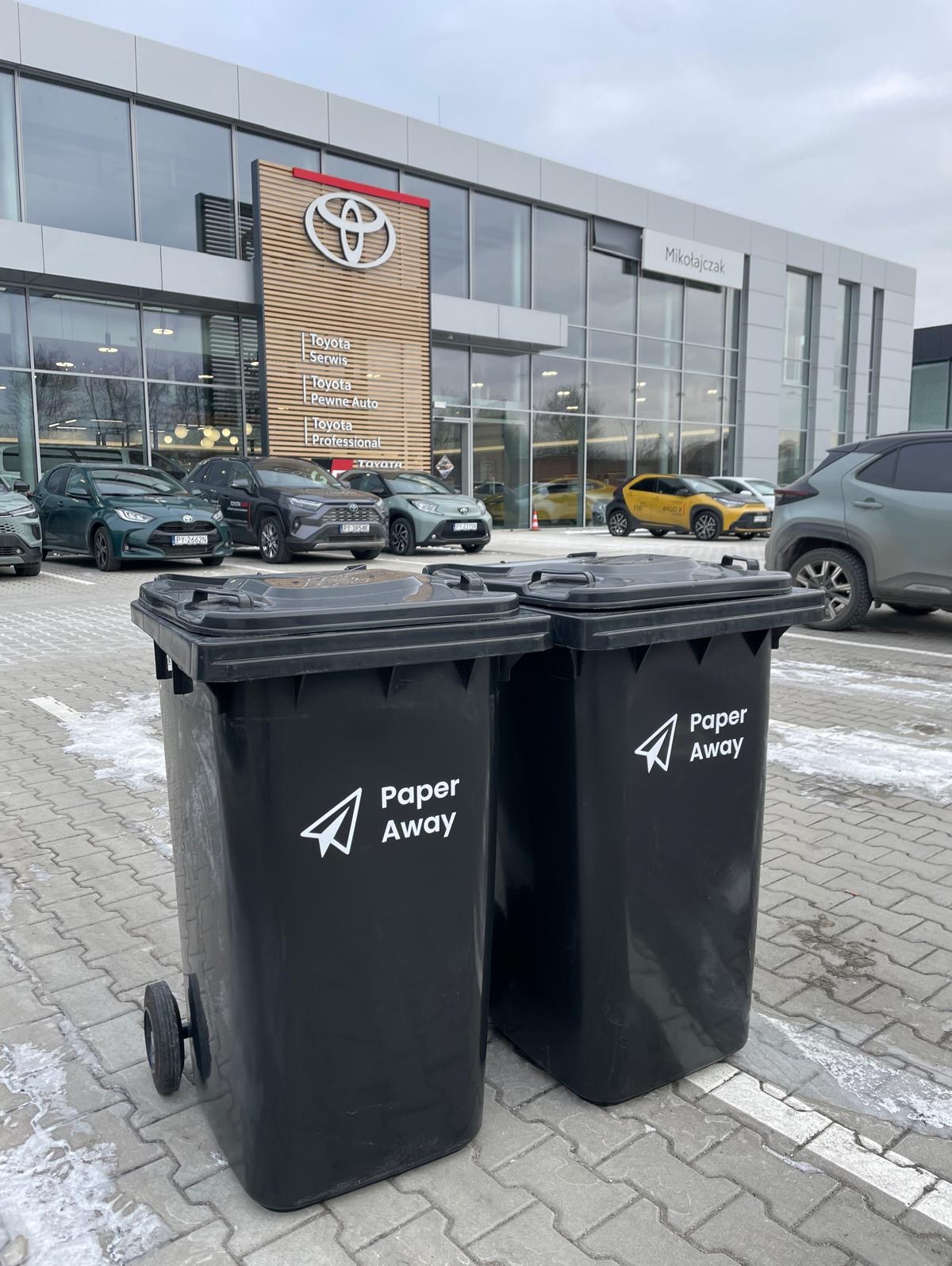 Dwa czarne pojemniki na kółkach z logo Paper Away stoją przed salonem samochodowym Toyota Mikołajczak. Usługa odbioru dokumentów.