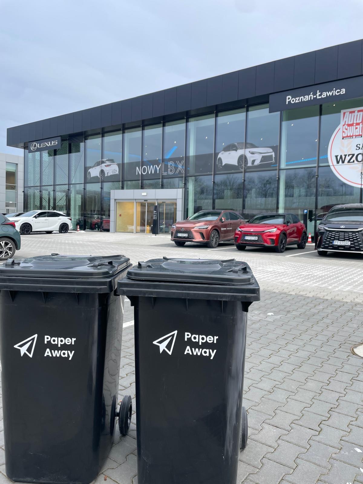 Dwa czarne pojemniki na kółkach z logo Paper Away na tle salonu Lexus Poznań-Ławica i zaparkowanych aut. Odbiór dokumentów do niszczenia.