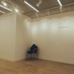 Puste, jasne pomieszczenie z panelami podłogowymi, krzesłem z kurtką w rogu i widocznymi instalacjami elektrycznymi na ścianie.