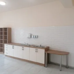 Jasne pomieszczenie z umywalkami w zabudowie, białe kafelki na ścianie, regał z półkami i stolik na metalowych nogach.