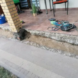 Przygotowanie podłoża pod taras z kostki brukowej, widoczne narzędzia budowlane i fragment kolumny z cegły klinkierowej
