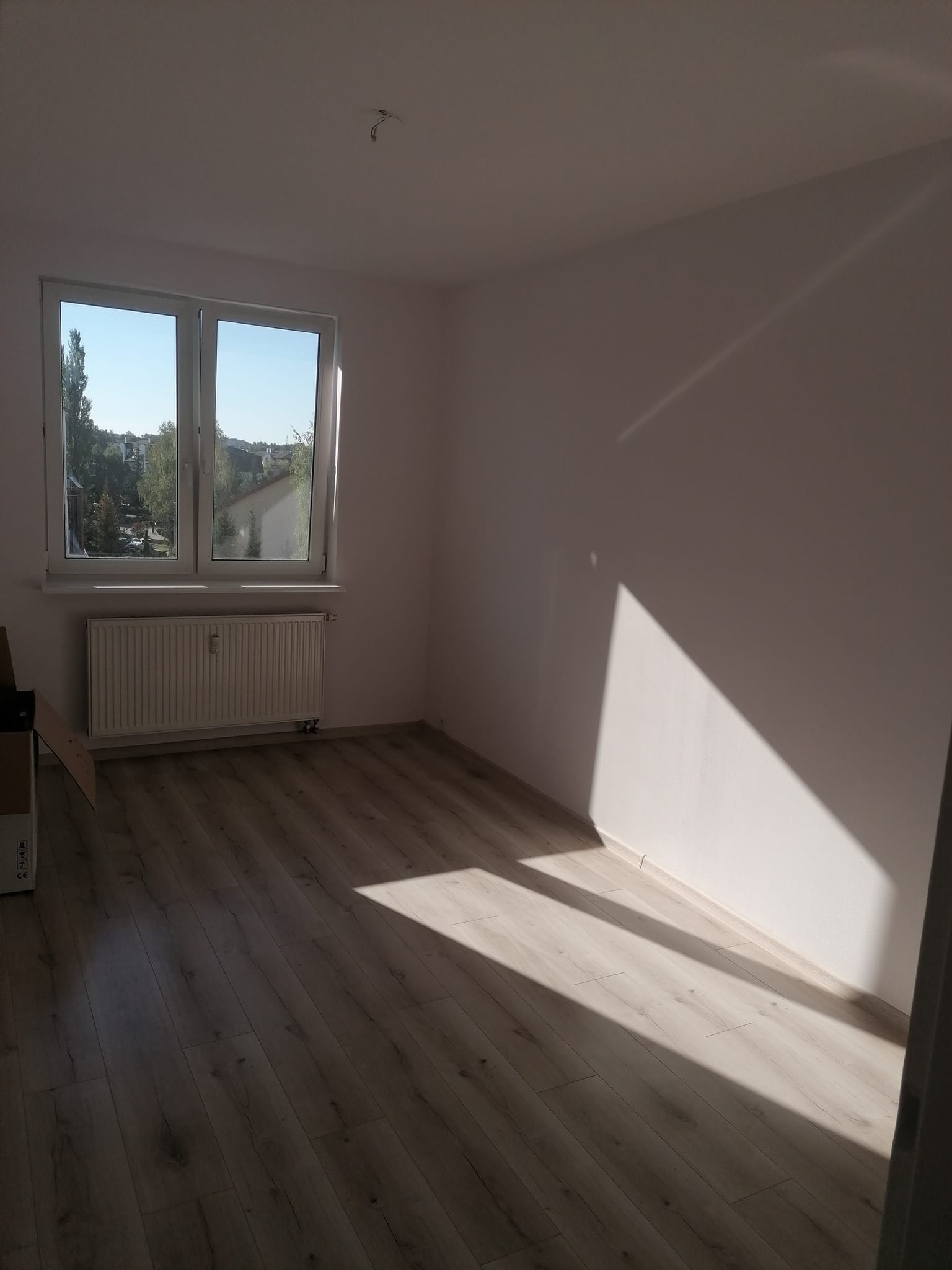 Pusty pokój z jasnymi panelami podłogowymi, białymi ścianami i grzejnikiem pod oknem, oświetlony popołudniowym słońcem tworzącym geometryczne cienie na ścianie.