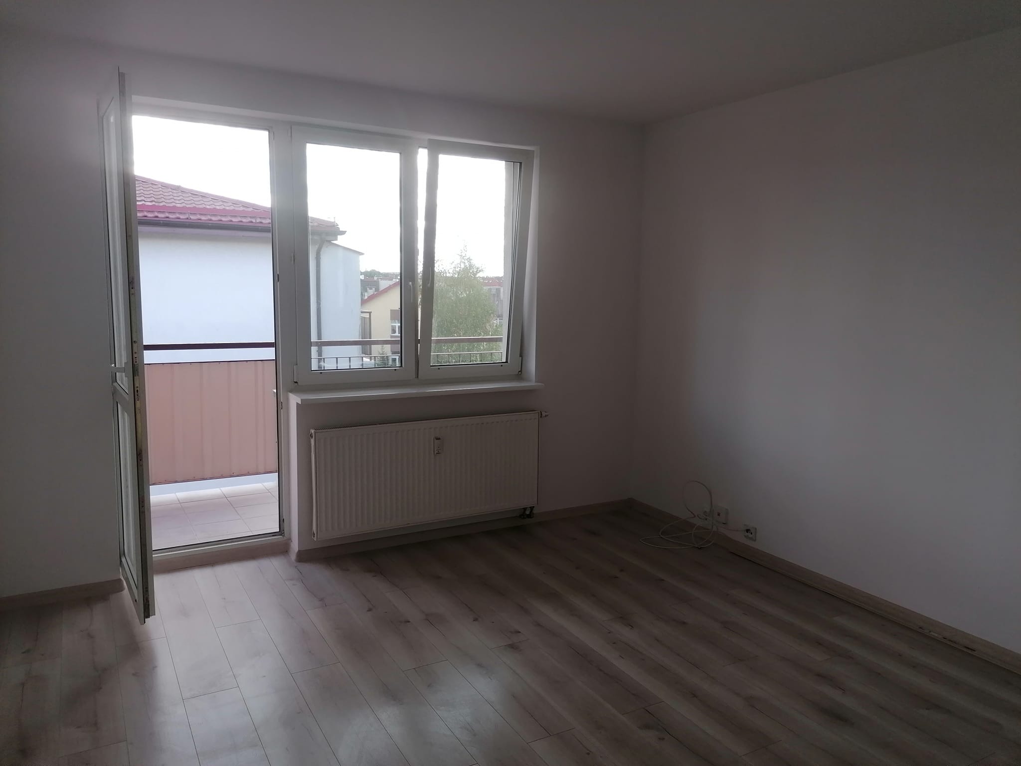 Pusty pokój z panelami podłogowymi w odcieniach szarości, otwartym balkonem z widokiem na budynek z czerwoną dachówką, białymi ścianami i grzejnikiem pod oknem.