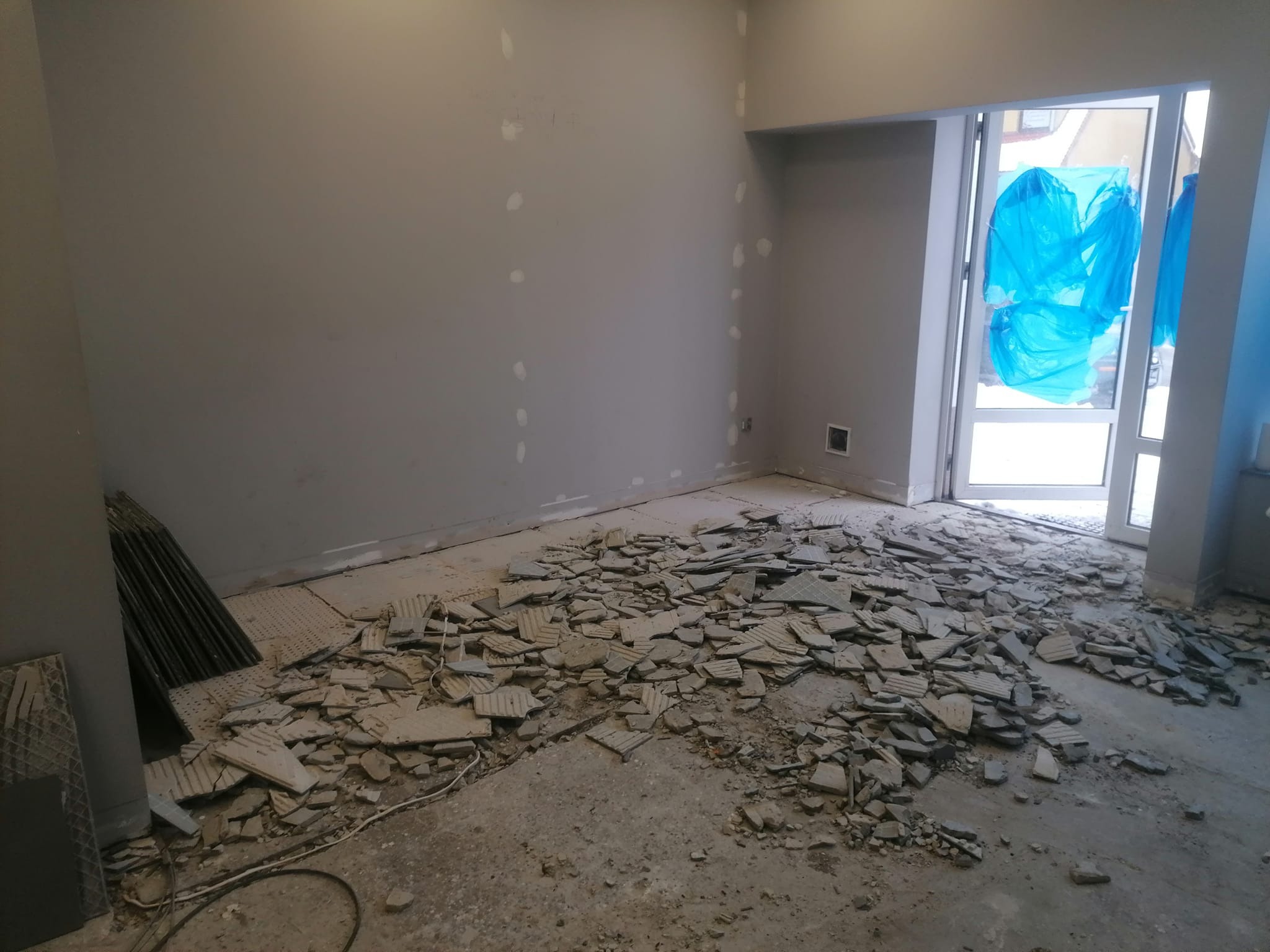 Pokój w trakcie remontu z fragmentami potłuczonych płytek na podłodze, ściany szare z widocznymi punktami po szpachlowaniu, okno zabezpieczone folią. W rogu pomieszczenia ułożone płytki. Widoczny...