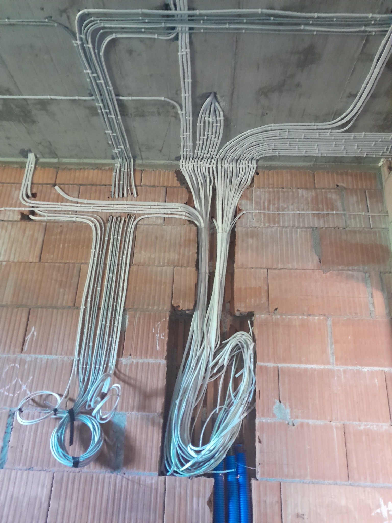 Rozprowadzenie przewodów elektrycznych w peszlach na surowym murze z cegły. Widoczne kable ułożone w korytach podtynkowych i na suficie. Etap prac instalacyjnych.