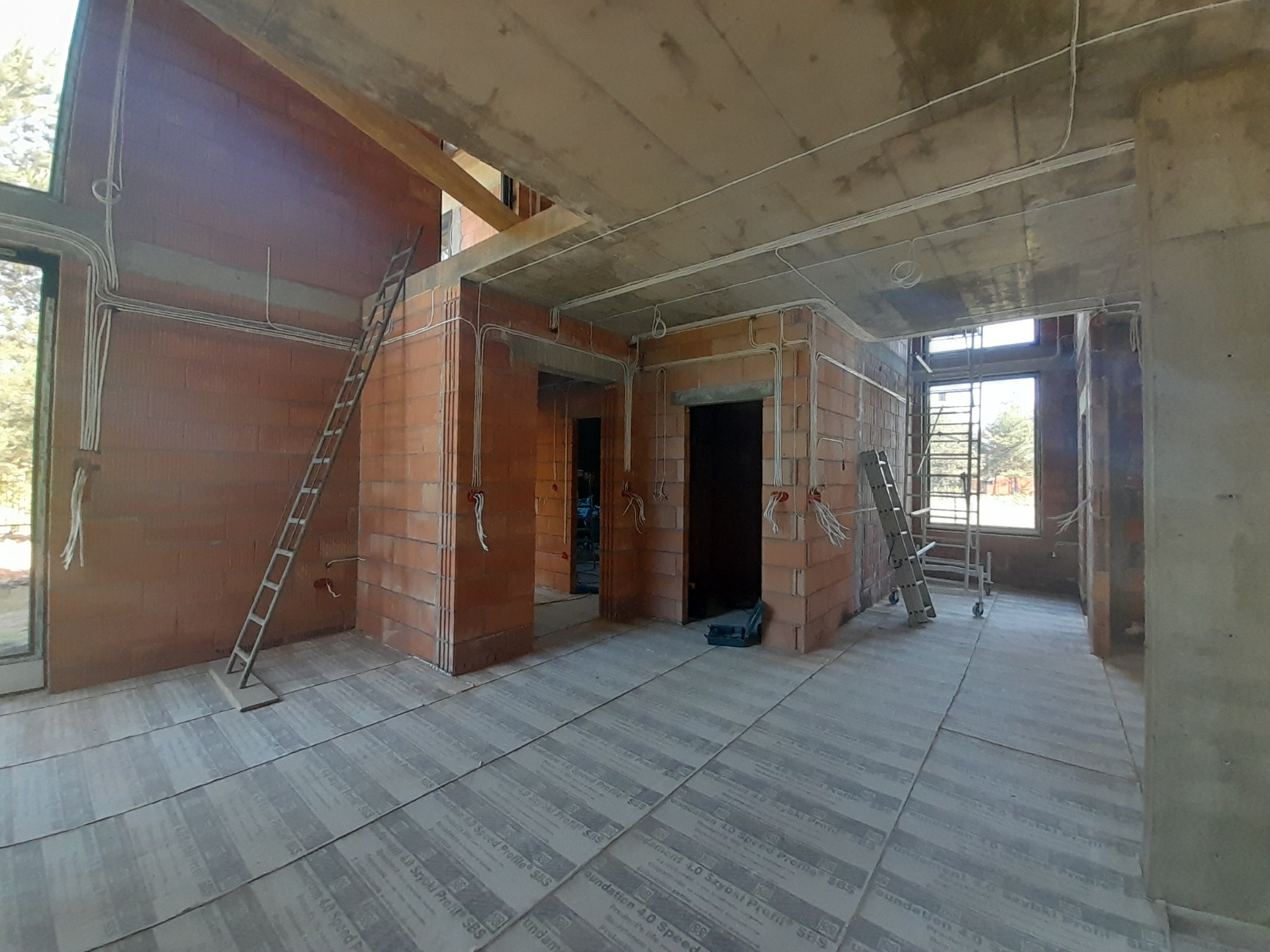 Wnętrze w trakcie budowy z widoczną instalacją elektryczną na ścianach z cegły i podciągniętymi kablami pod sufitem. Na podłodze płyty izolacyjne.