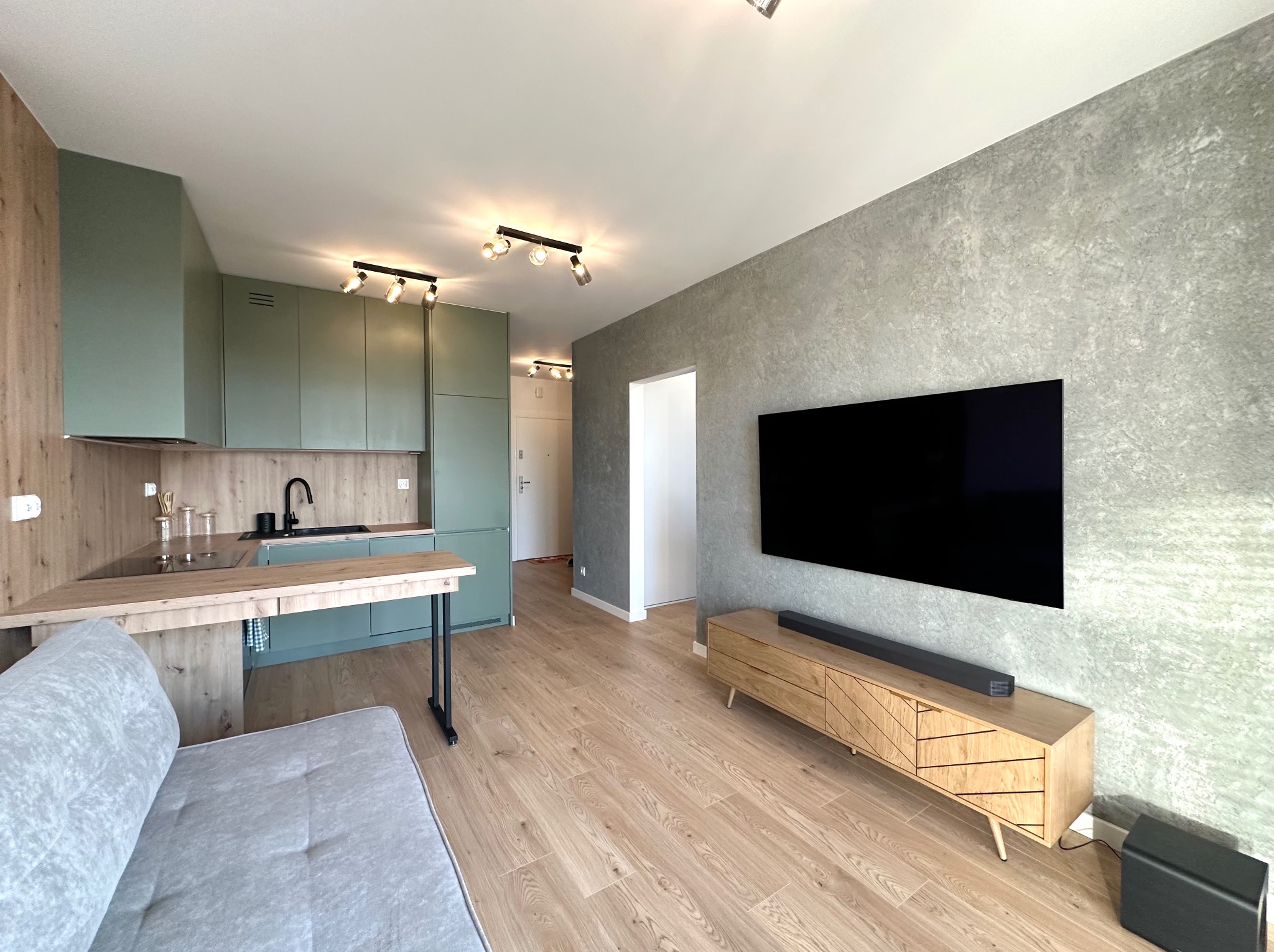 Nowoczesne wnętrze: kuchnia z zielonymi szafkami, drewniany blat, szara ściana z TV, drewniana podłoga i szara sofa. Oświetlenie punktowe.