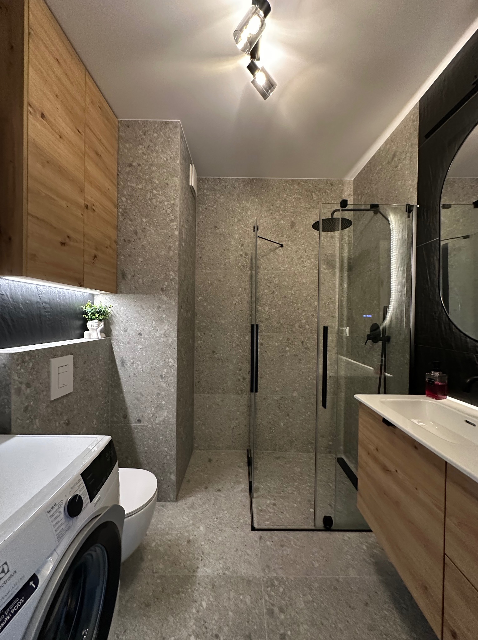 Nowoczesna łazienka z kabiną prysznicową typu walk-in, drewnianymi szafkami, szarą mozaiką na ścianach i podłodze oraz designerską lampą sufitową.