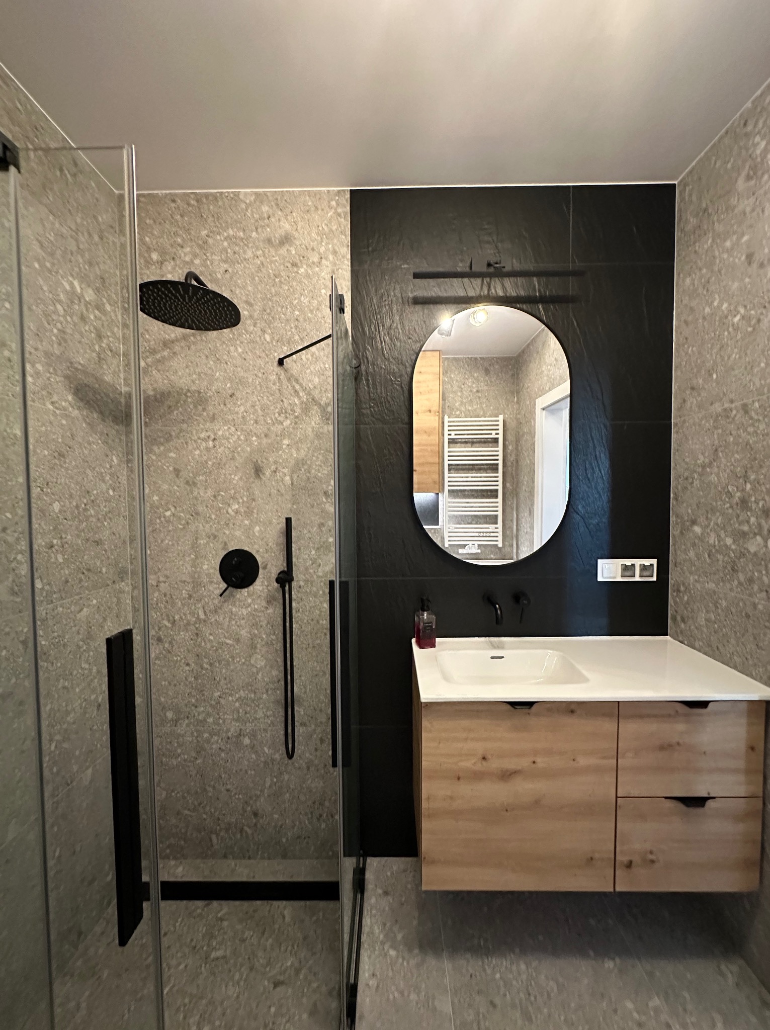 Nowoczesna łazienka z kabiną prysznicową typu walk-in, owalnym lustrem i drewnianą szafką pod umywalką. Czarna armatura i szare płytki tworzą elegancki kontrast.