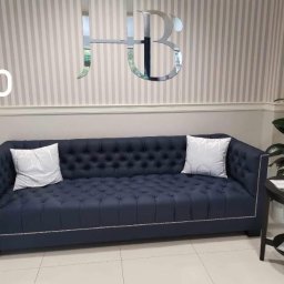 Dar Pikowania Natalia Kokoszka - Granatowa sofa Chesterfield z poduszkami na tle ściany w paski i lustrzanego logo HB. Sofa z ozdobnymi ćwiekami, elegancki design.