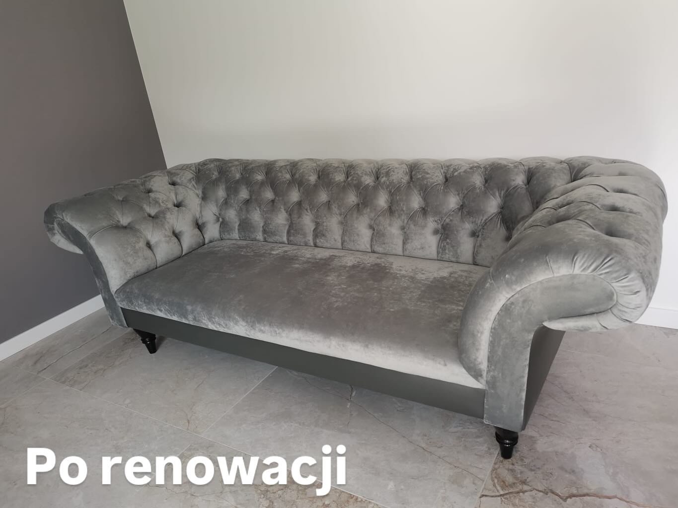 Szara sofa typu chesterfield po renowacji, z głębokimi pikowaniami i eleganckimi podłokietnikami, ustawiona na tle ściany w dwóch odcieniach szarości.
