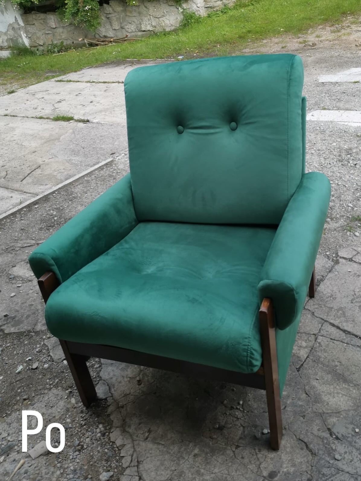 Szmaragdowy fotel z drewnianymi nogami, obity miękkim welurem z guzikami, stojący na zewnątrz na tle betonowej nawierzchni. Widoczny napis 'Po'.