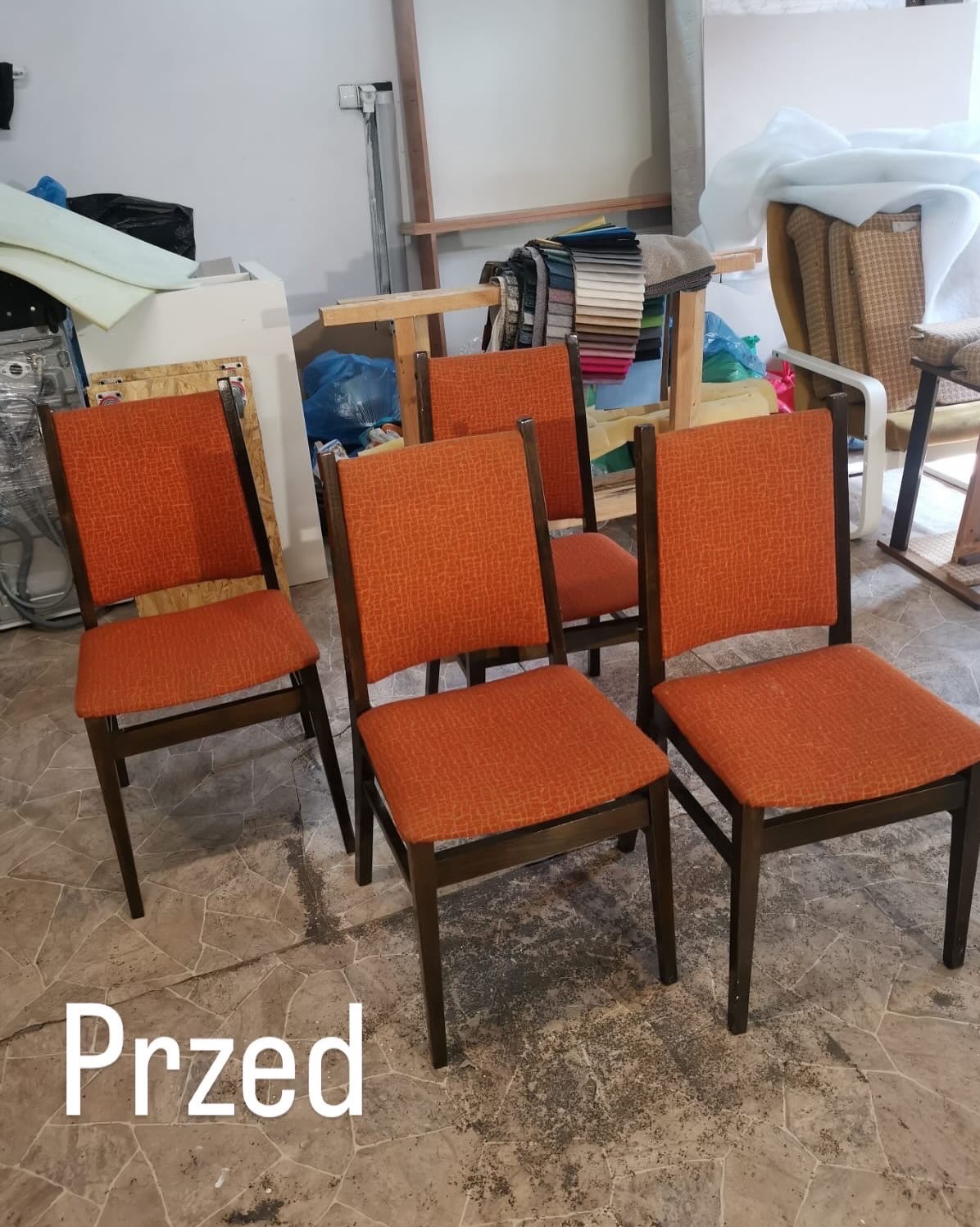 Cztery krzesła z pomarańczową tapicerką w warsztacie tapicerskim, widoczne materiały i narzędzia. Stan 'przed' renowacją, z bliska, na tle podłogi z wzorem.