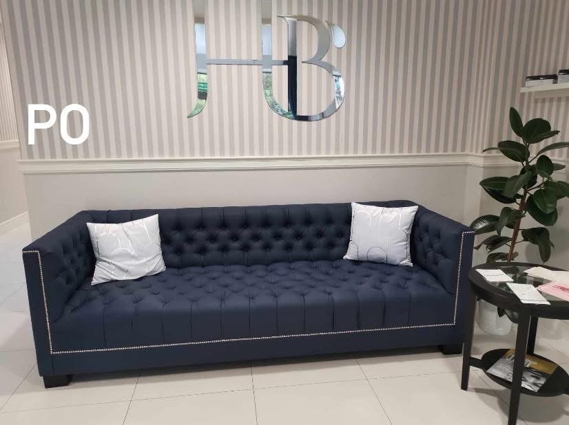 Granatowa sofa Chesterfield z poduszkami na tle ściany w paski i lustrzanego logo HB. Sofa z ozdobnymi ćwiekami, elegancki design.