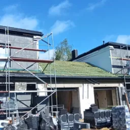 Budowa domu z rusztowaniami, częściowo pokrytym zieloną dachówką, z oknami zabezpieczonymi folią i zaparkowanym samochodem budowlanym na pierwszym planie.