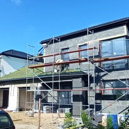 MAŁSTEF Stefan Kozeł - Budowa domu: ocieplony budynek w szarym tynku, okna zabezpieczone folią, rusztowania, zielony dach, częściowo widoczny pracownik na dachu. Słoneczny dzień.