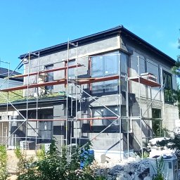 MAŁSTEF Stefan Kozeł - Dwukondygnacyjny dom w trakcie ocieplania, z widocznymi rusztowaniami i częściowo zamontowanymi oknami. Prace budowlane w słoneczny dzień.