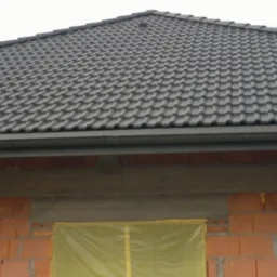 Częściowy widok dachu z szarej dachówki ceramicznej nad murem z czerwonej cegły. Okno zabezpieczone folią. Widoczne rynny.
