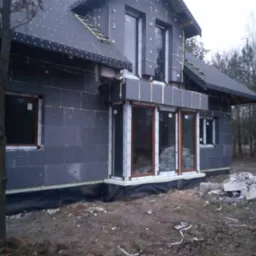 Dom murowany w trakcie budowy, ściany ocieplone styropianem grafitowym, widoczne okna z brązowymi ramami i stos bloczków Ytong na ziemi.