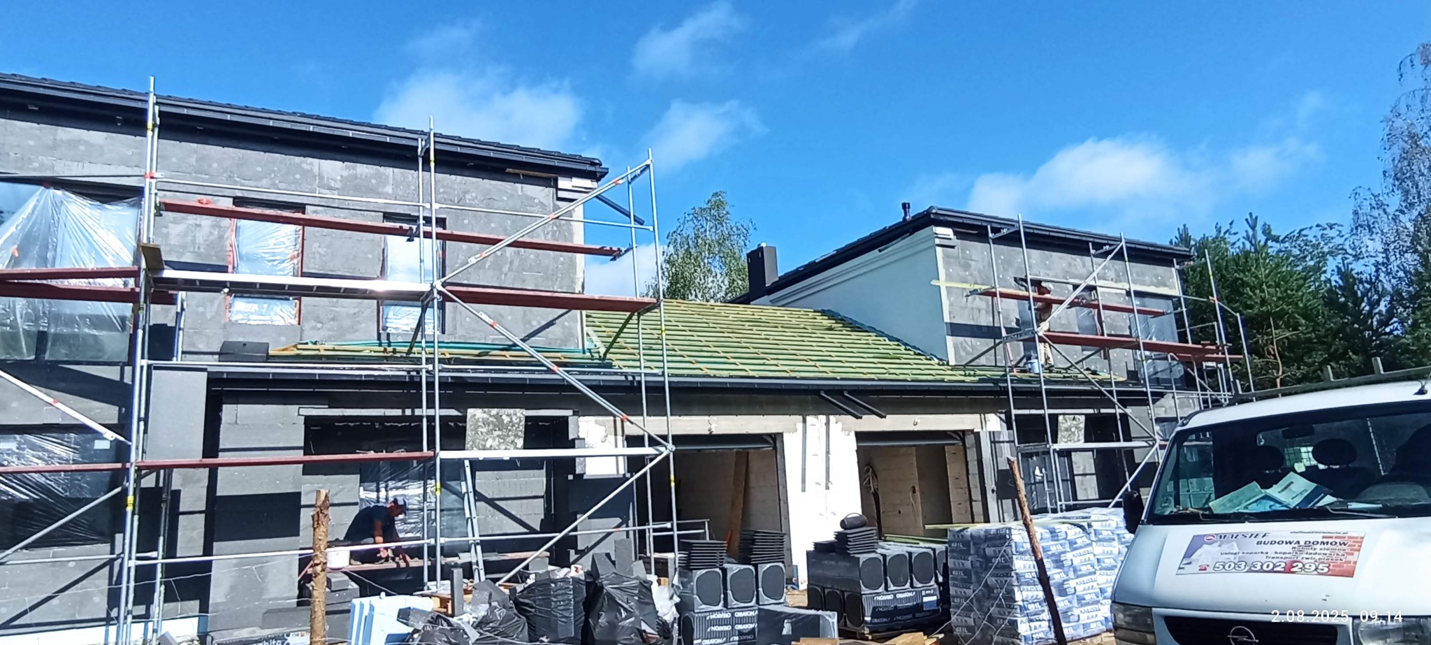 Budowa domu z rusztowaniami, częściowo pokrytym zieloną dachówką, z oknami zabezpieczonymi folią i zaparkowanym samochodem budowlanym na pierwszym planie.