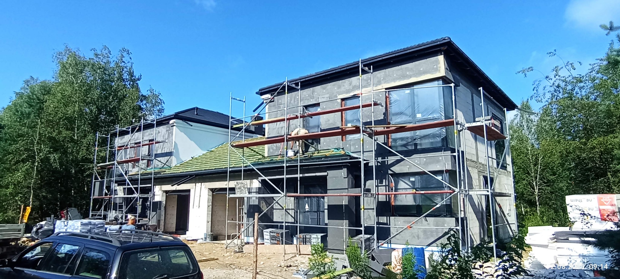 Budowa domu: ocieplony budynek w szarym tynku, okna zabezpieczone folią, rusztowania, zielony dach, częściowo widoczny pracownik na dachu. Słoneczny dzień.