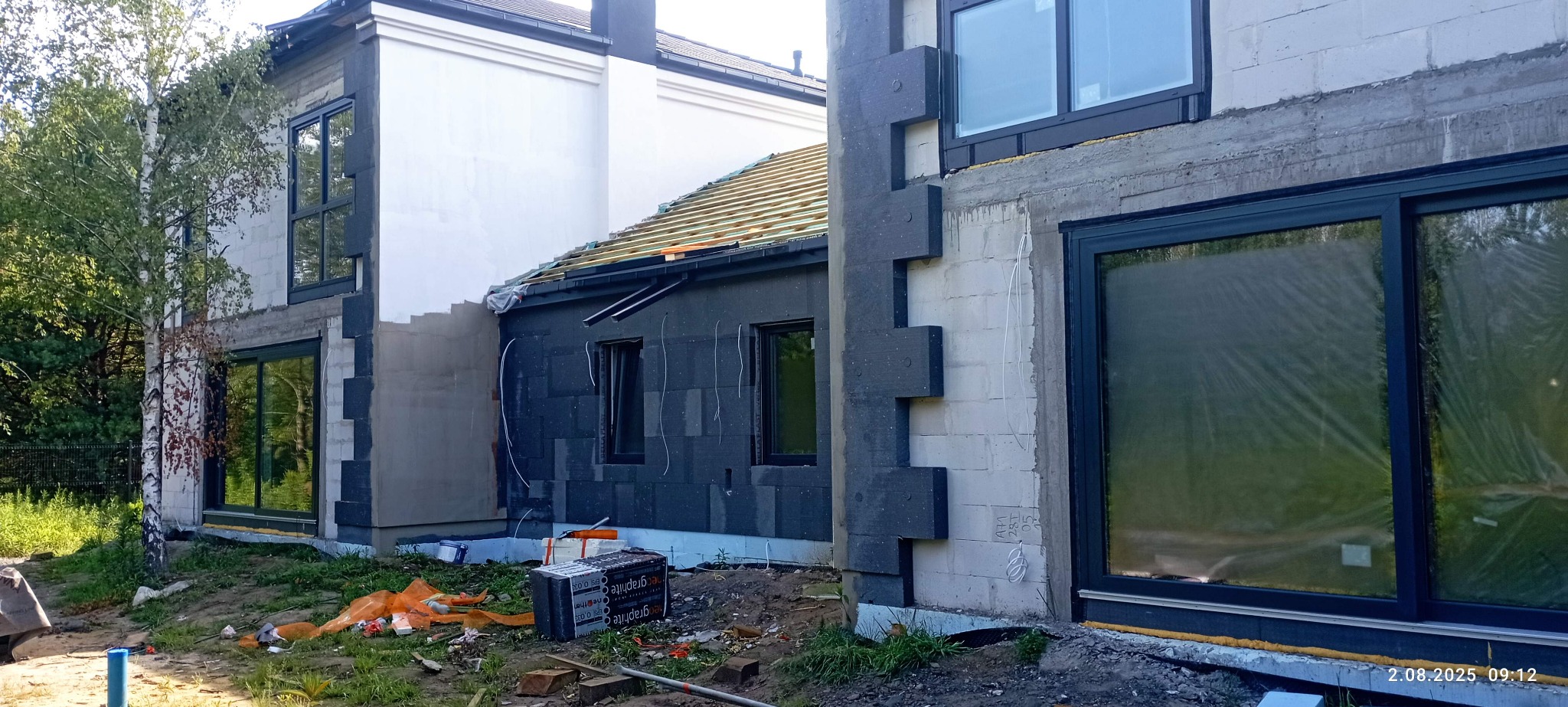 Budowa domu w trakcie: ocieplenie ścian grafitowym styropianem, widoczne okna z folią ochronną, więźba dachowa, białe bloczki. Prace budowlane w toku.