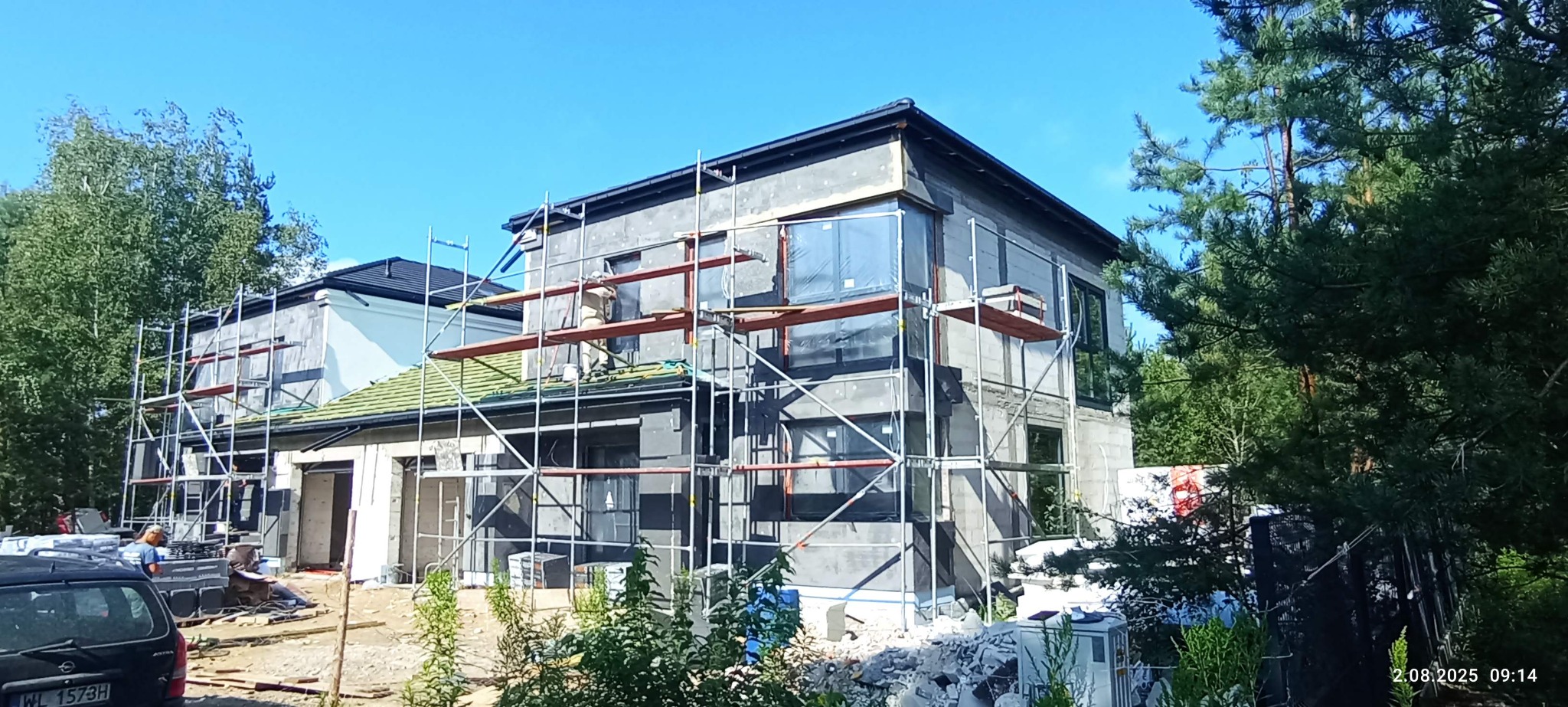 Dwukondygnacyjny dom w trakcie ocieplania, z widocznymi rusztowaniami i częściowo zamontowanymi oknami. Prace budowlane w słoneczny dzień.