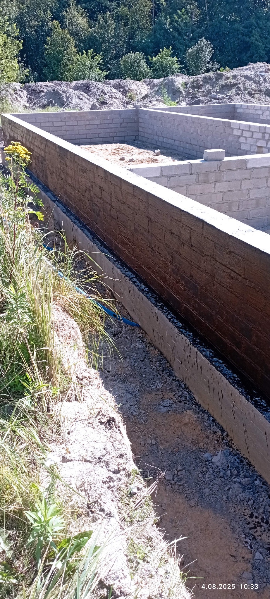 Zabezpieczone fundamenty z bloczków betonowych, izolowane czarną substancją, widoczne trawy i ziemia. Budowa w toku, z bliska, w słoneczny dzień.