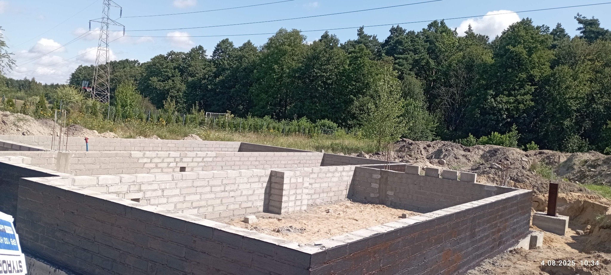 Wzniesione mury fundamentowe z bloczków betonowych, zabezpieczone izolacją pionową. W tle zieleń drzew i słup wysokiego napięcia. Widok z góry.