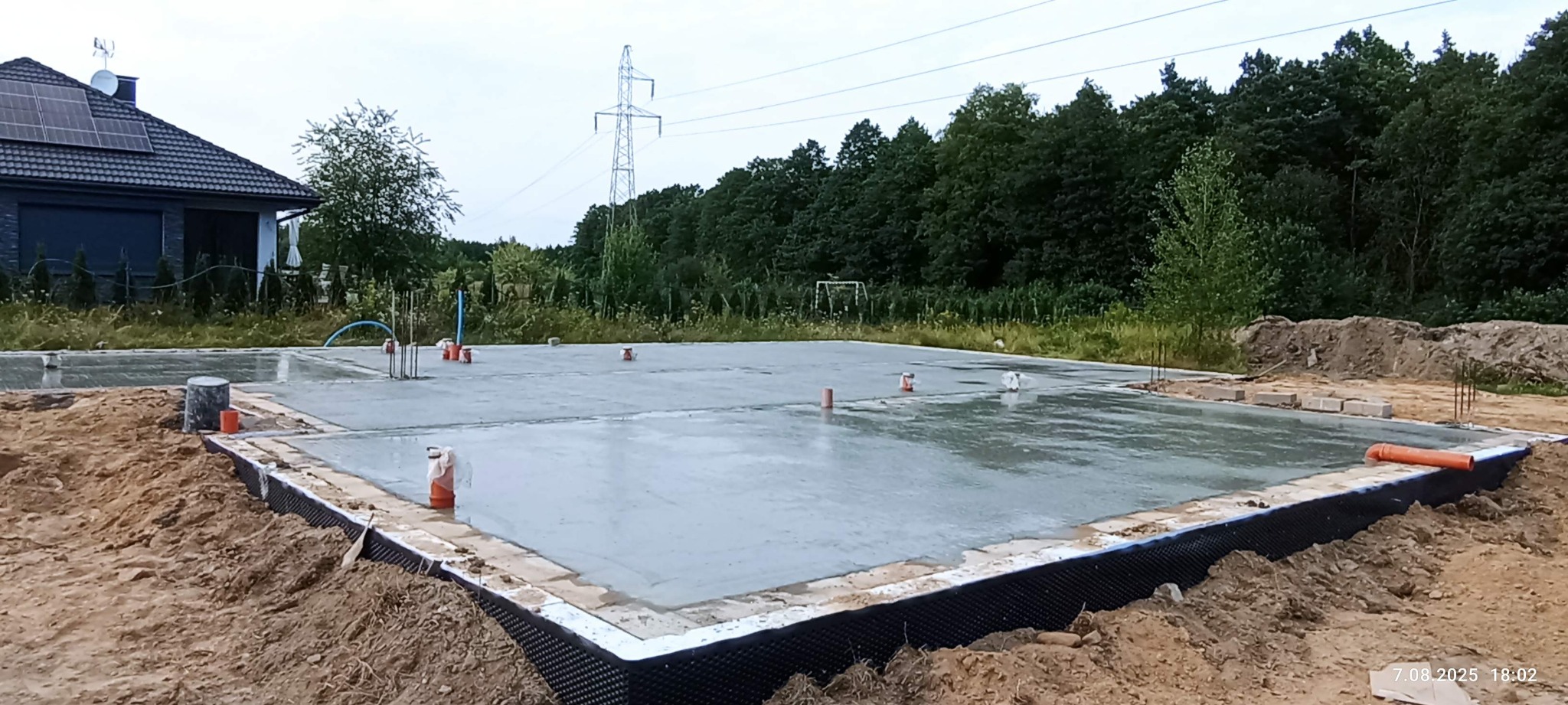 Zalany fundament domu jednorodzinnego z hydroizolacją, widoczne wystające rury instalacyjne i zbrojenia. W tle dom z panelami solarnymi i linia wysokiego napięcia.