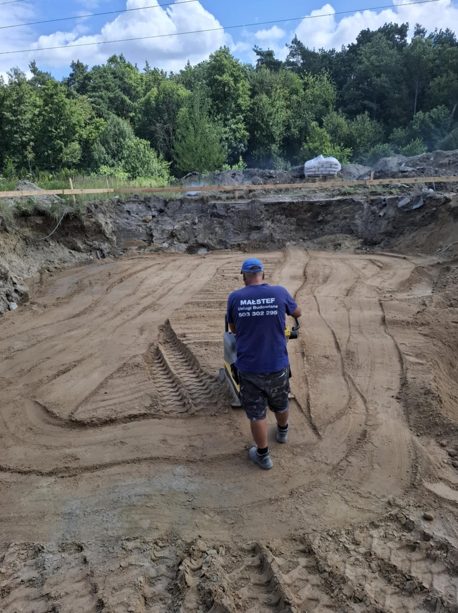 Pracownik zagęszcza piasek w wykopie pod budowę. Widoczne ślady po zagęszczarce, teren wyrównany, w tle drzewa i worki z materiałem budowlanym.