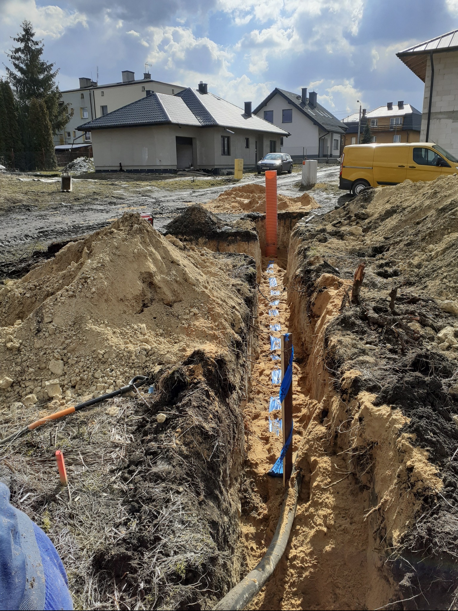 Wykop z ułożonymi rurami (pomarańczowa karbowana i czarna) zabezpieczonymi niebieską folią. W tle zabudowa jednorodzinna i żółty samochód dostawczy.