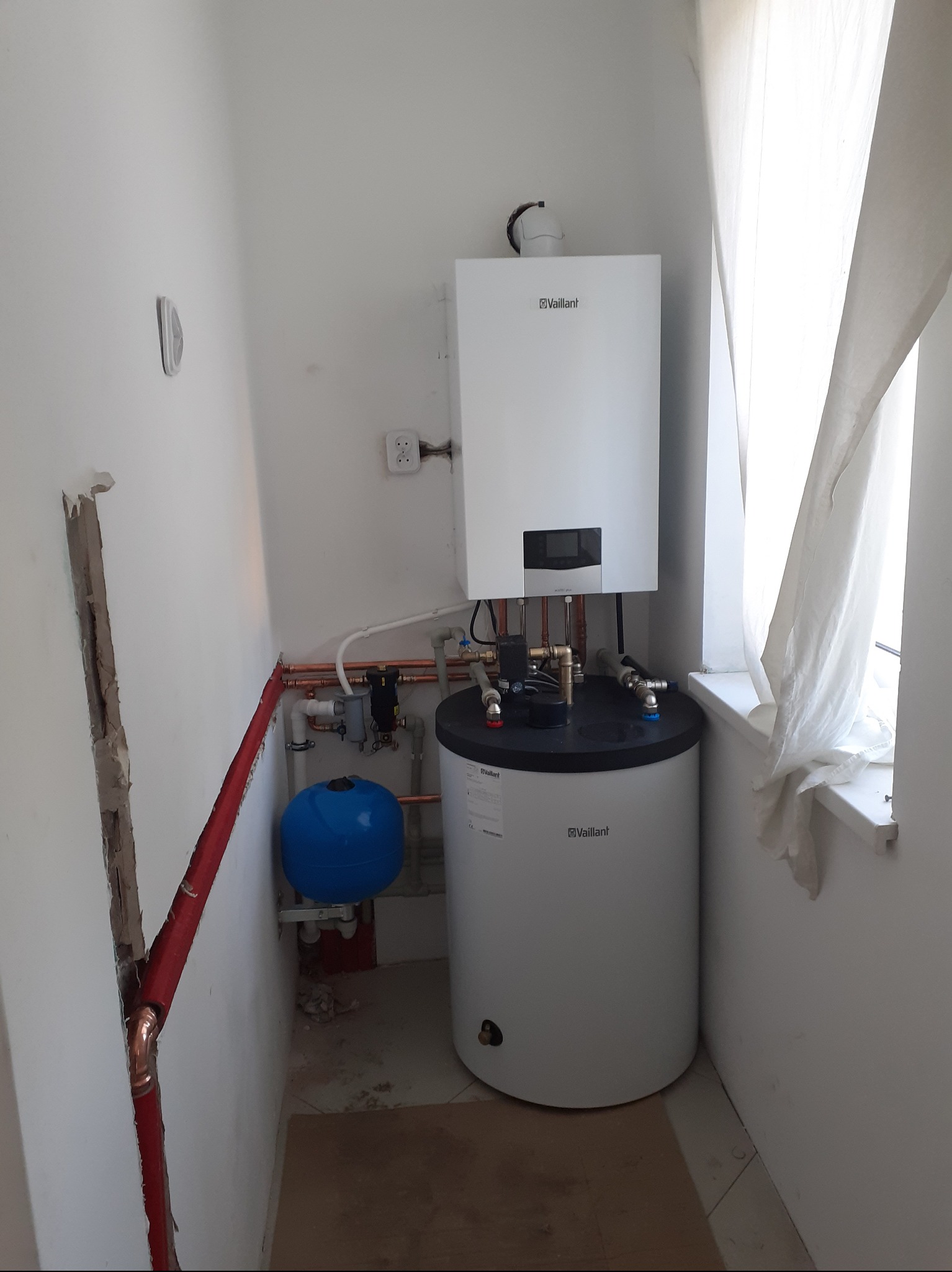 Biały piec gazowy Vaillant z zasobnikiem, instalacja z miedzianych rur, niebieskie naczynie przeponowe w małej kotłowni z oknem i zasłoną.