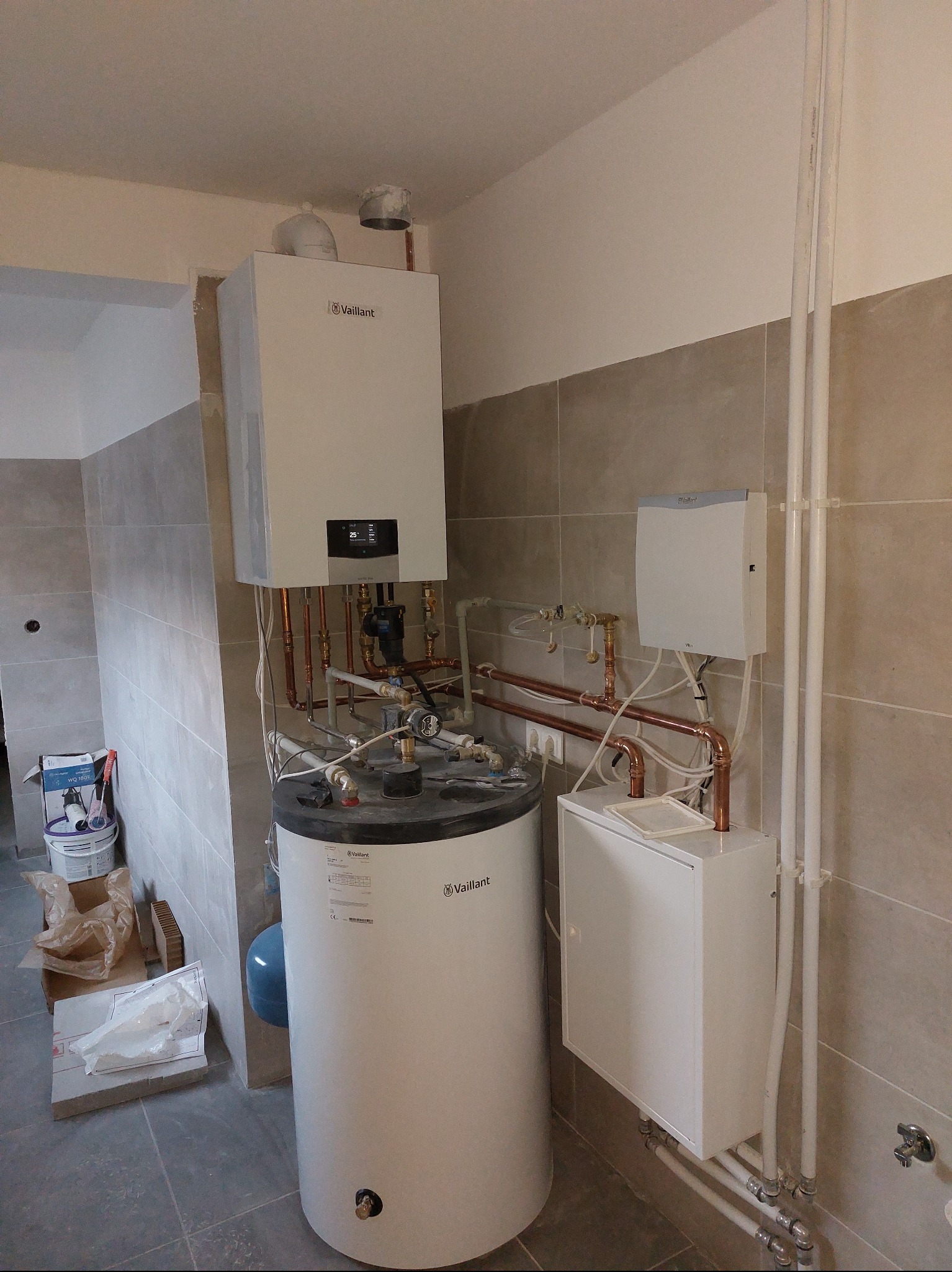 Biały piec gazowy Vaillant z zasobnikiem i osprzętem, zamontowany w pomieszczeniu z szarymi płytkami. Widoczne miedziane rury i białe plastikowe rurki.