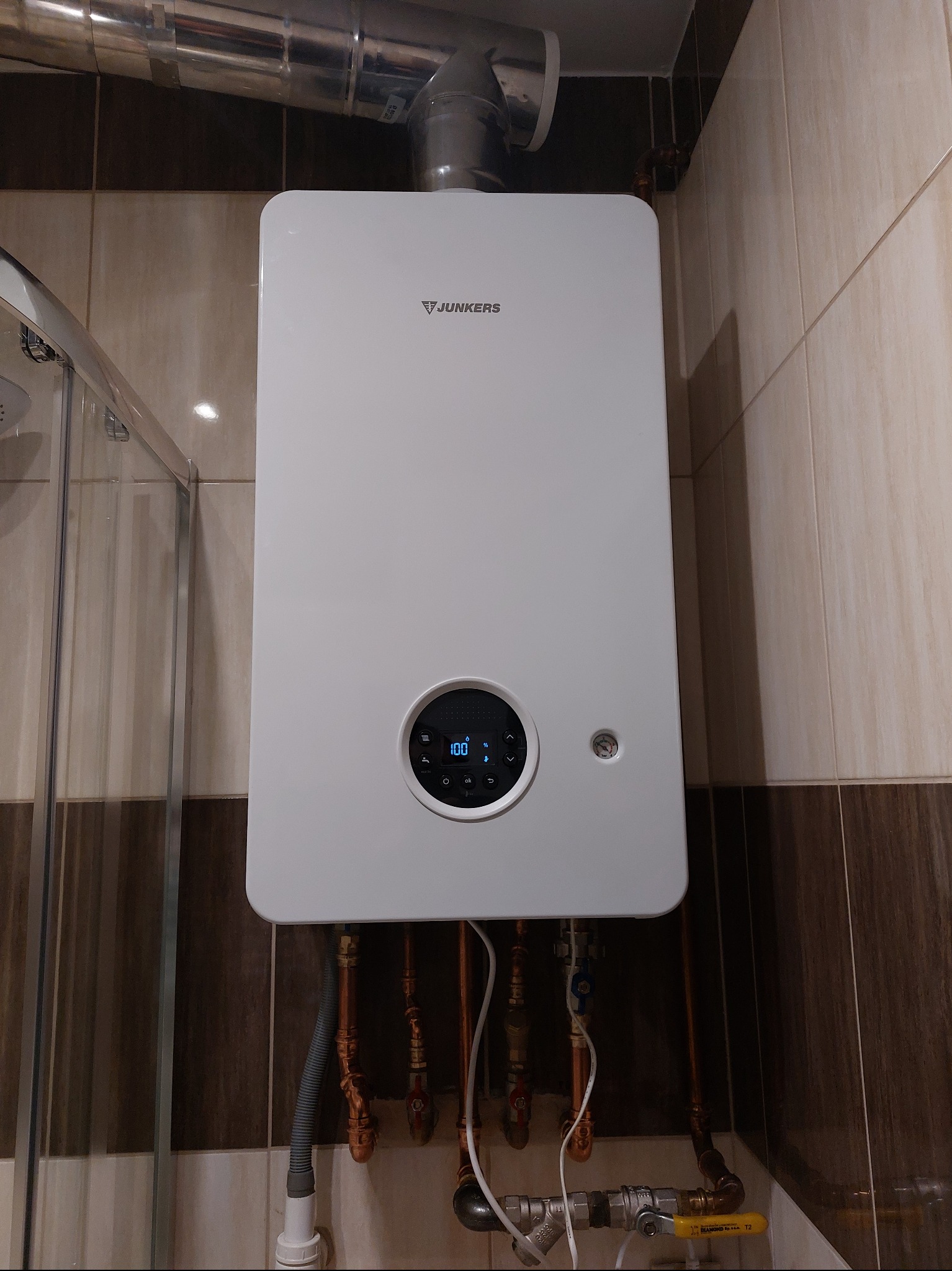 Biały piec gazowy Junkers zamontowany na ścianie z beżowo-brązowymi płytkami, widoczne rury doprowadzające media i fragment kabiny prysznicowej po lewej stronie.