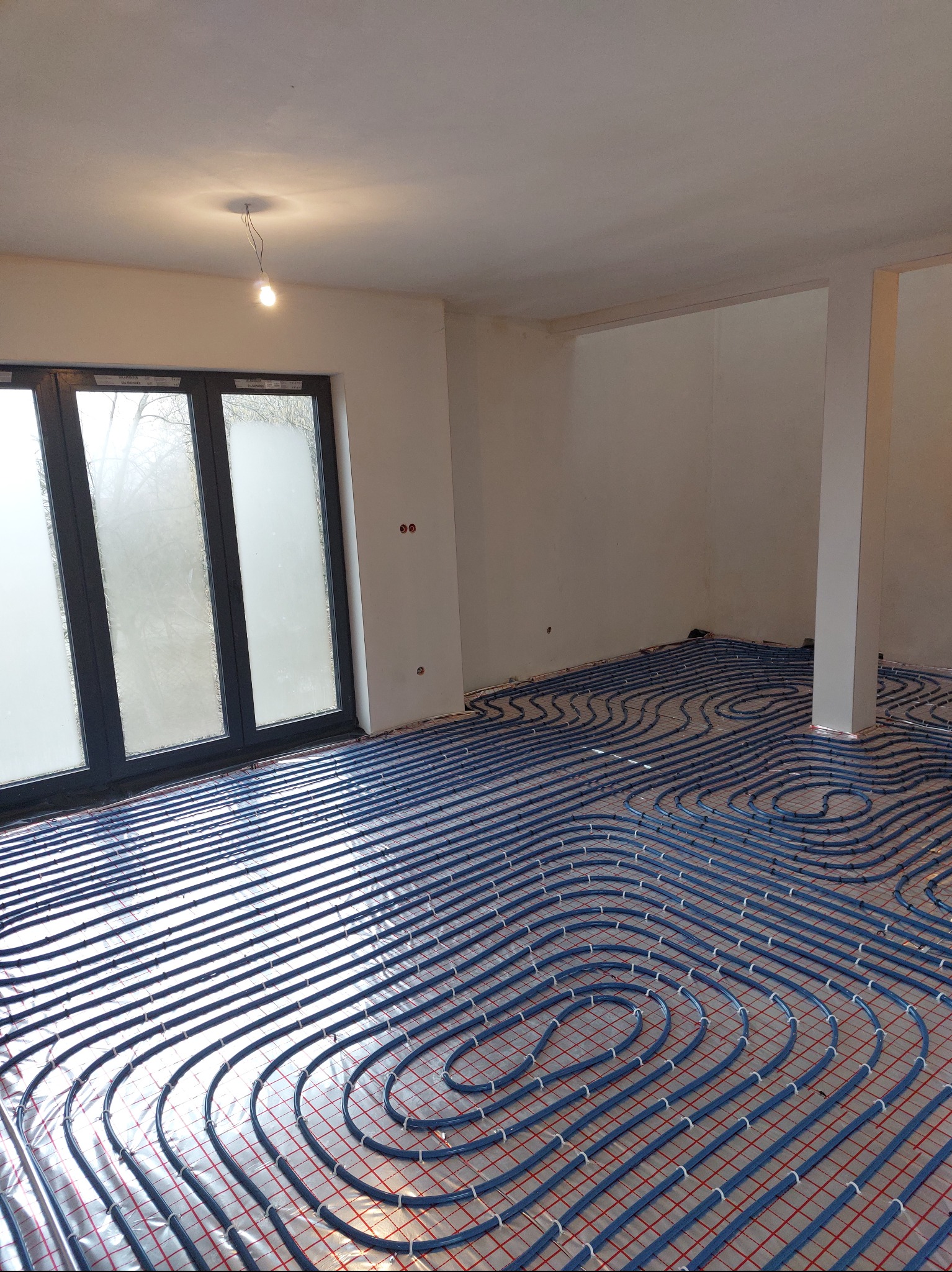 Niebieskie rury ogrzewania podłogowego ułożone spiralnie na srebrnej folii z czerwoną siatką w surowym wnętrzu z oknem i wiszącą żarówką. Etap instalacji przed wylewką.