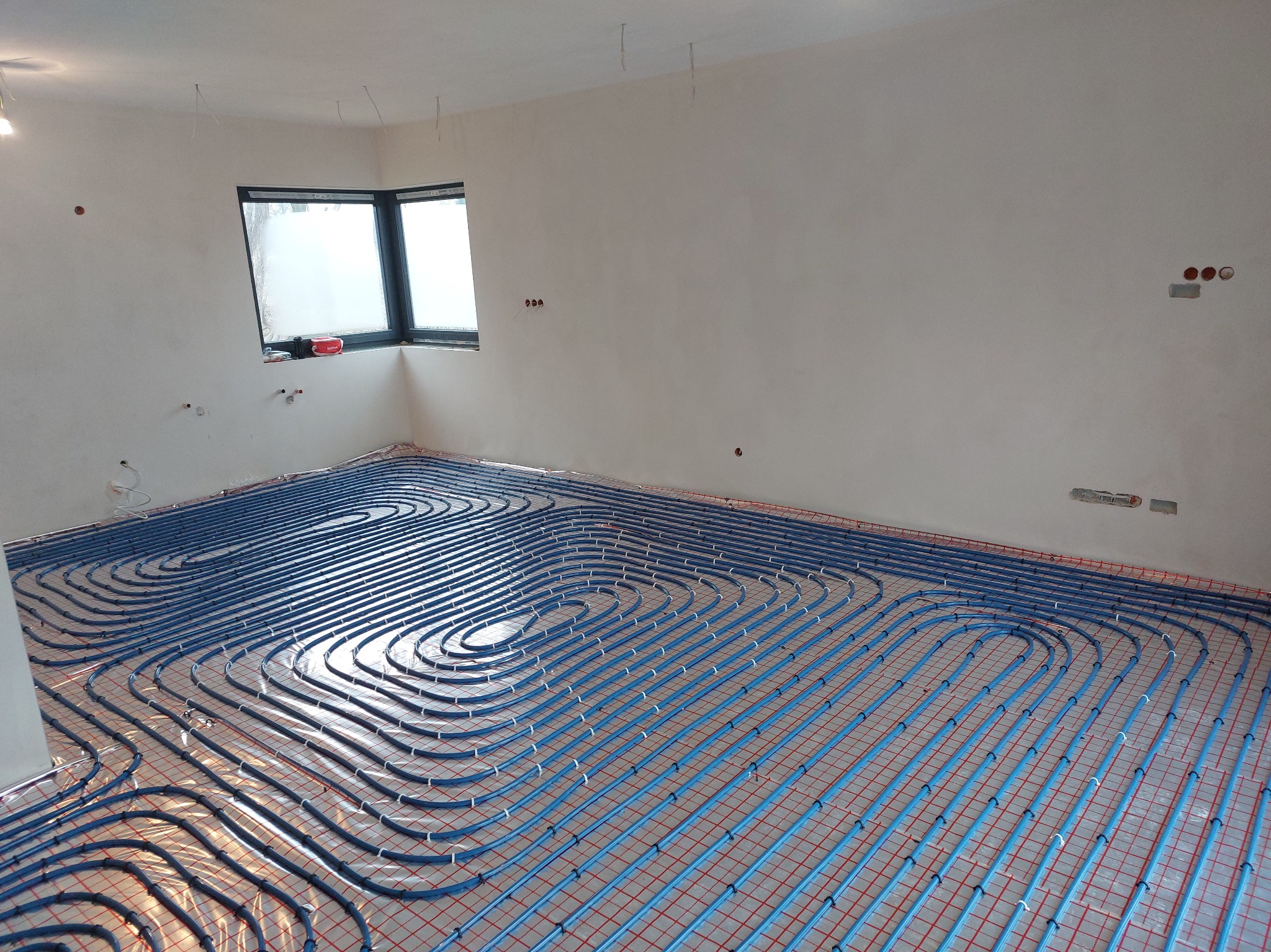 Niebieskie rury ogrzewania podłogowego ułożone na czerwonej siatce w jasnym pokoju z oknem. Widoczne elementy instalacji na ścianach.