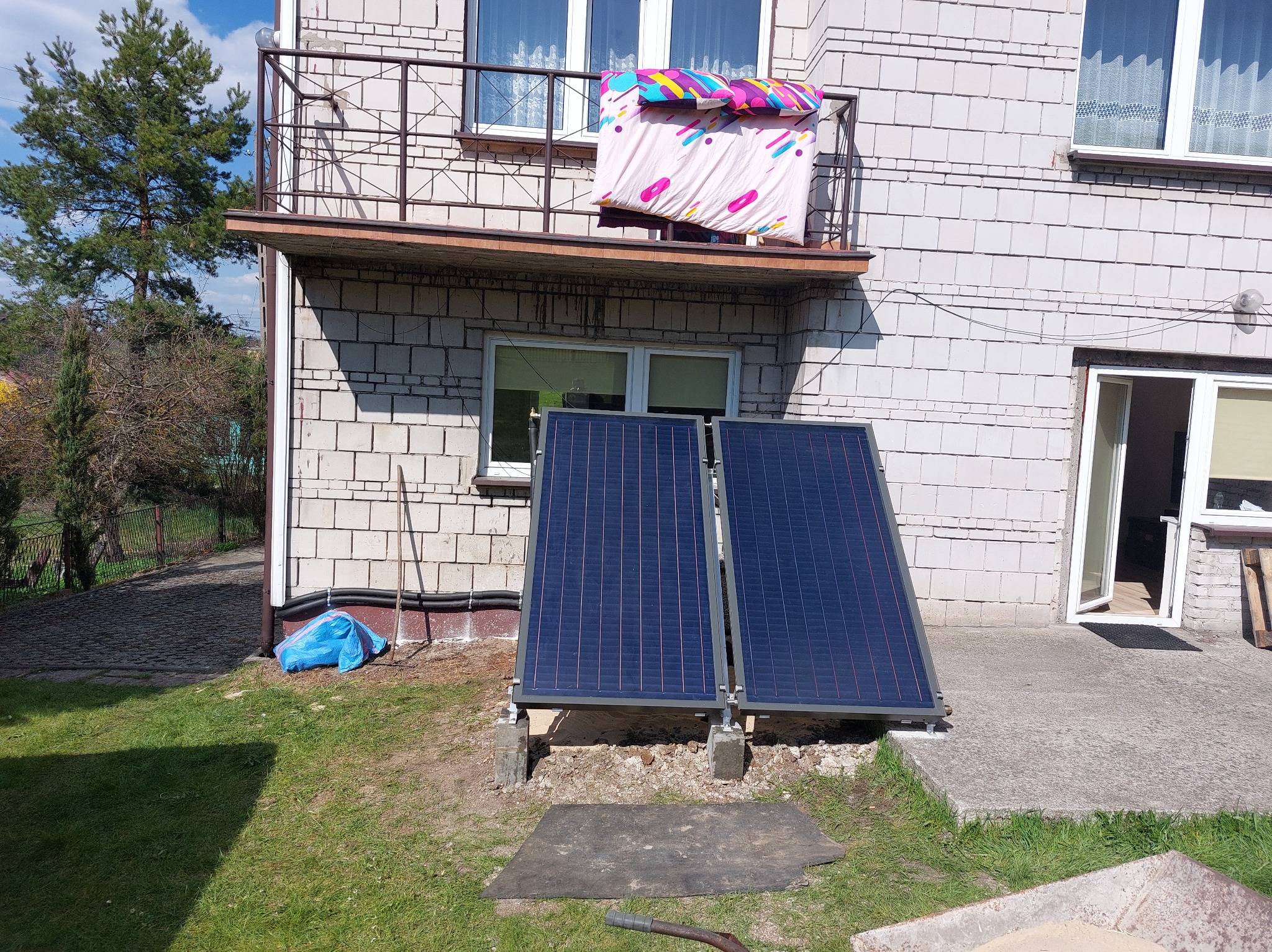 Dwa panele solarne zamontowane przy ścianie domu z balkonem, na trawniku. Widoczne elementy instalacji i otoczenie budynku. Ujęcie z poziomu gruntu.